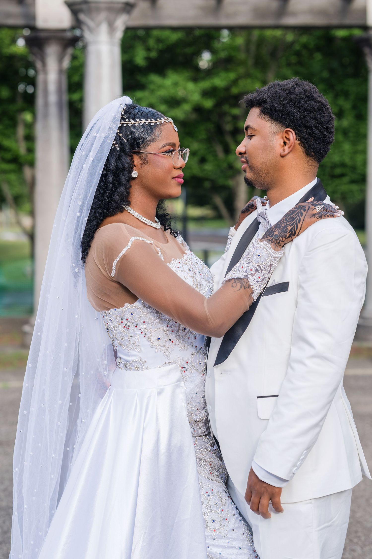A beautiful wedding couple embraces outdoors, with the bride in a white lace gown and veil and groom in a white tuxedo.