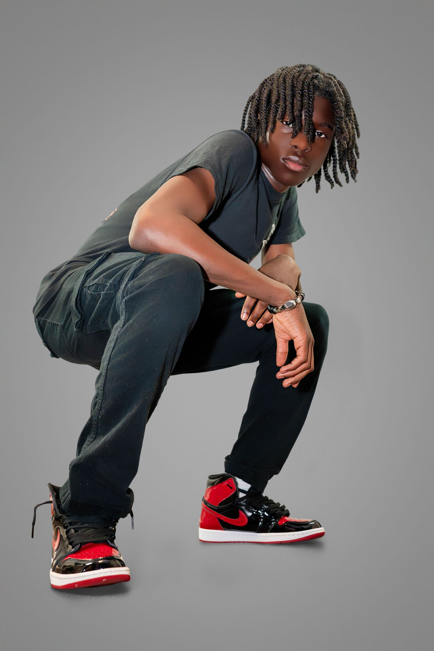 Stylish person in dark clothing and red sneakers crouches in dynamic pose against grey studio backdrop.
