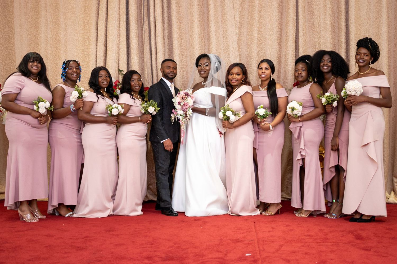 A wedding party poses together on a red carpet wearing elegant pink bridesmaid dresses and a white wedding gown.