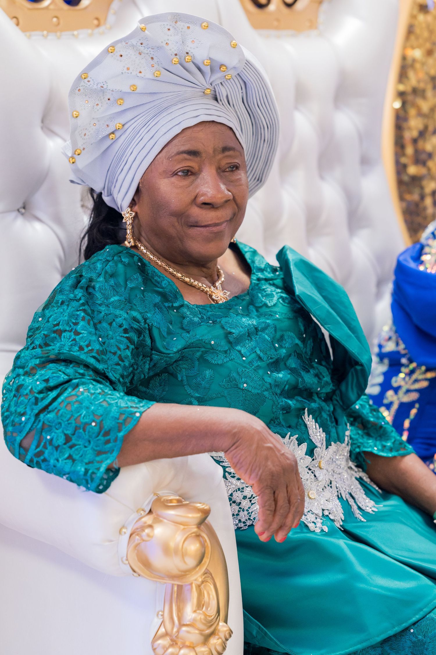 A woman wearing a teal lace dress and grey headwrap sits regally in a white throne-like chair.