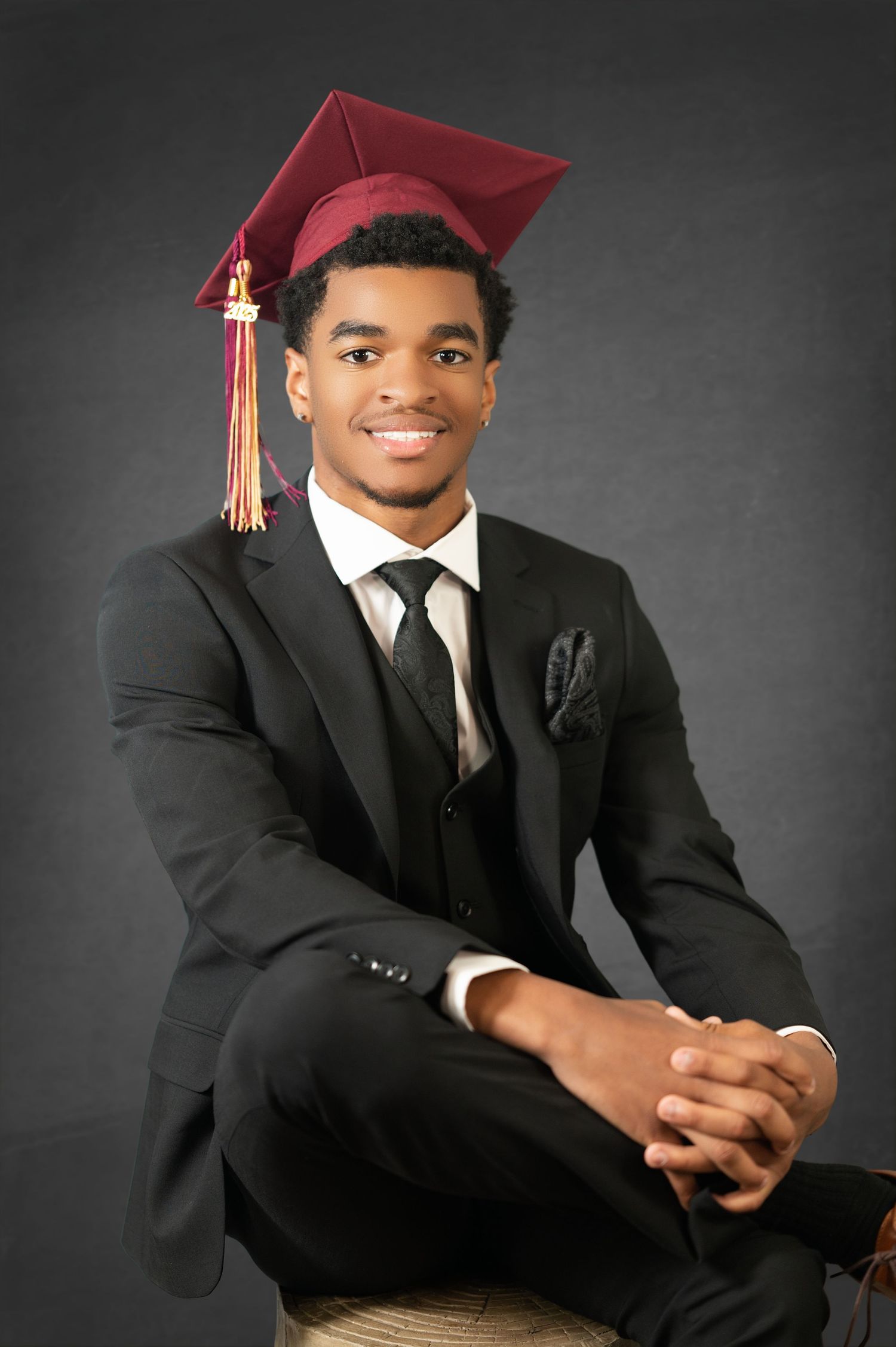 A graduate in a burgundy cap and black suit sits for a professional portrait against a dark grey background.