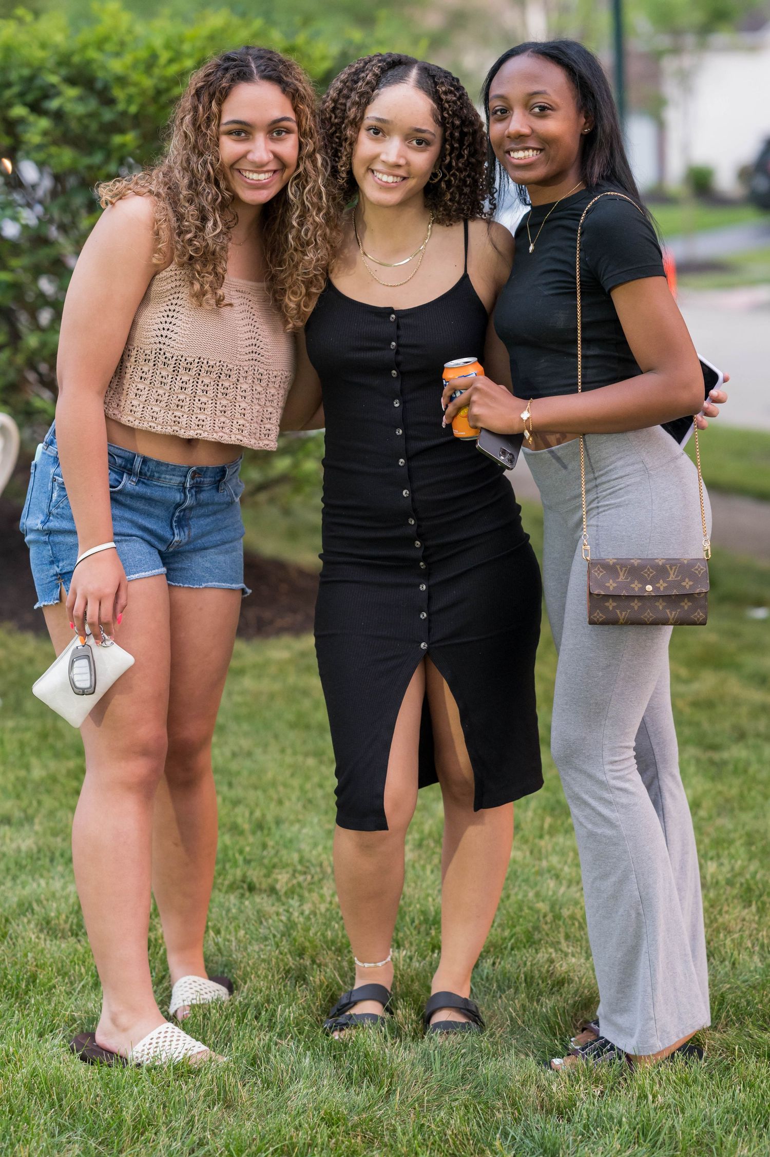 Three friends pose outdoors on grass, wearing stylish casual outfits including a black dress, denim shorts and grey pants.