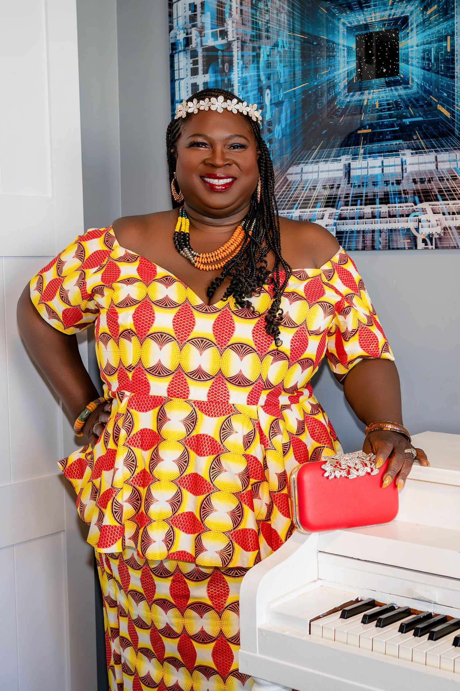 Elegant woman in vibrant yellow and red patterned dress with pearl tiara poses beside white piano.