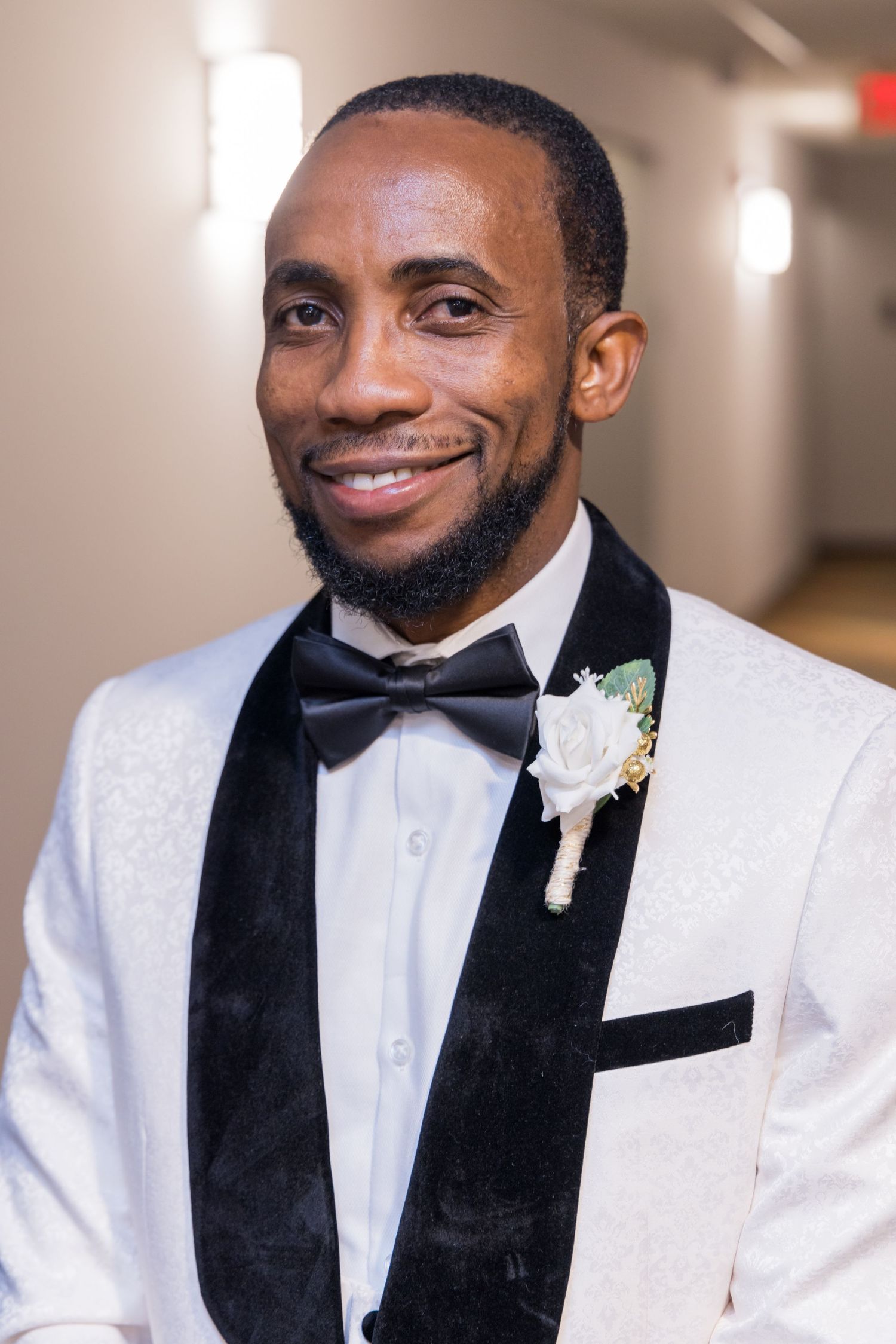 Man in white tuxedo jacket with black lapels and bow tie smiles while wearing a white boutonniere.