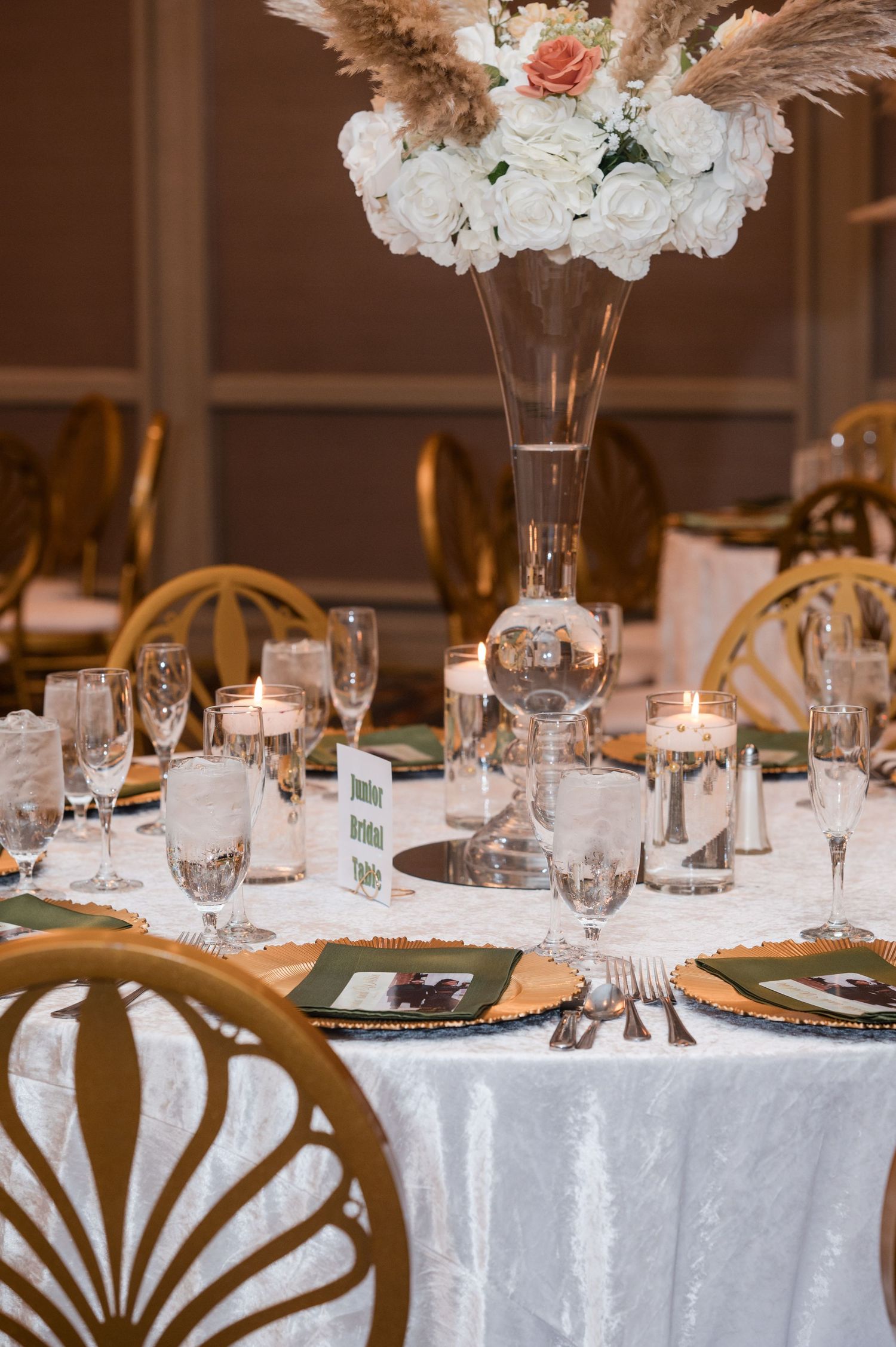 Elegant wedding reception table with white floral centerpiece, gold chairs, and glowing candles on white tablecloth.