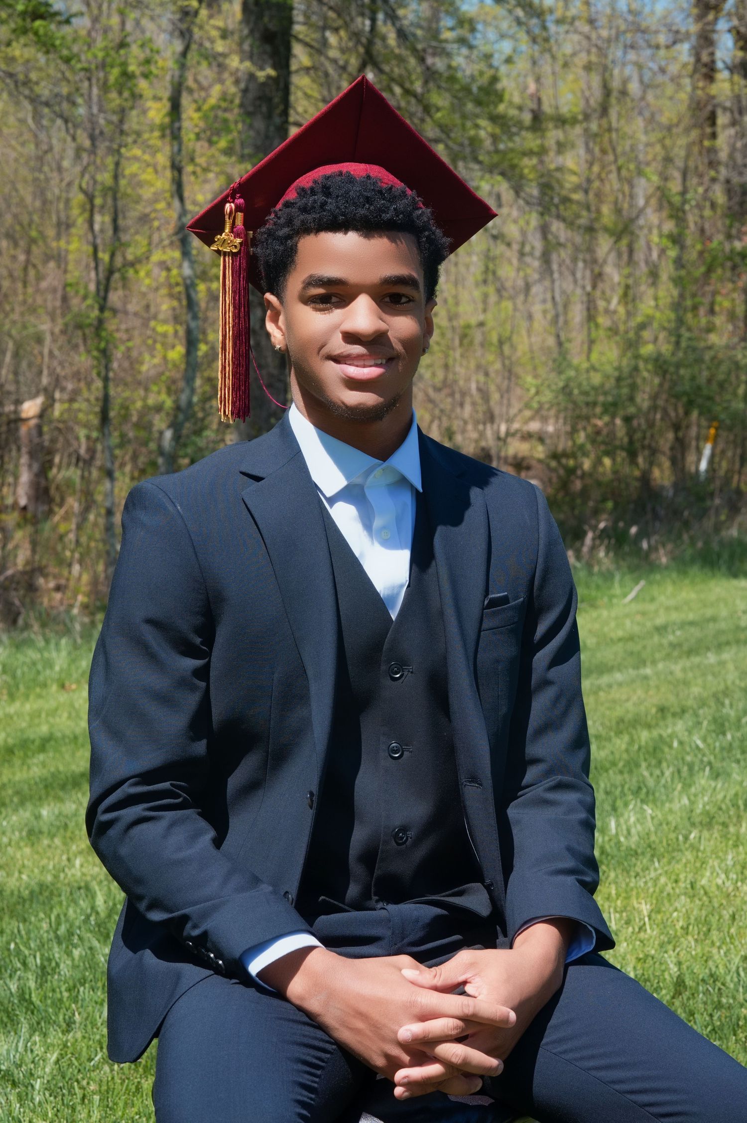 Graduate in maroon cap and navy suit posing outdoors for portrait on sunny spring day with trees in background.
