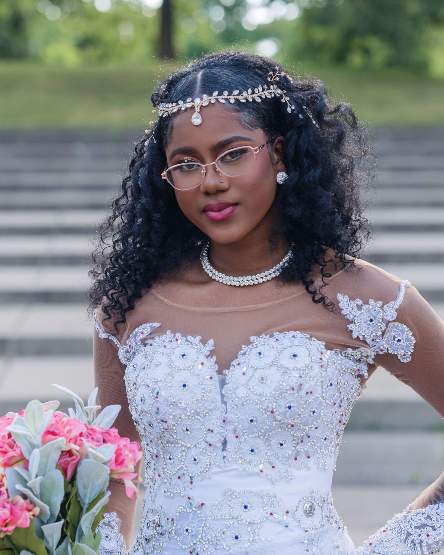 Beautiful bridal portrait featuring curly hair, crystal headpiece, sheer lace wedding gown and pink bouquet outdoors.