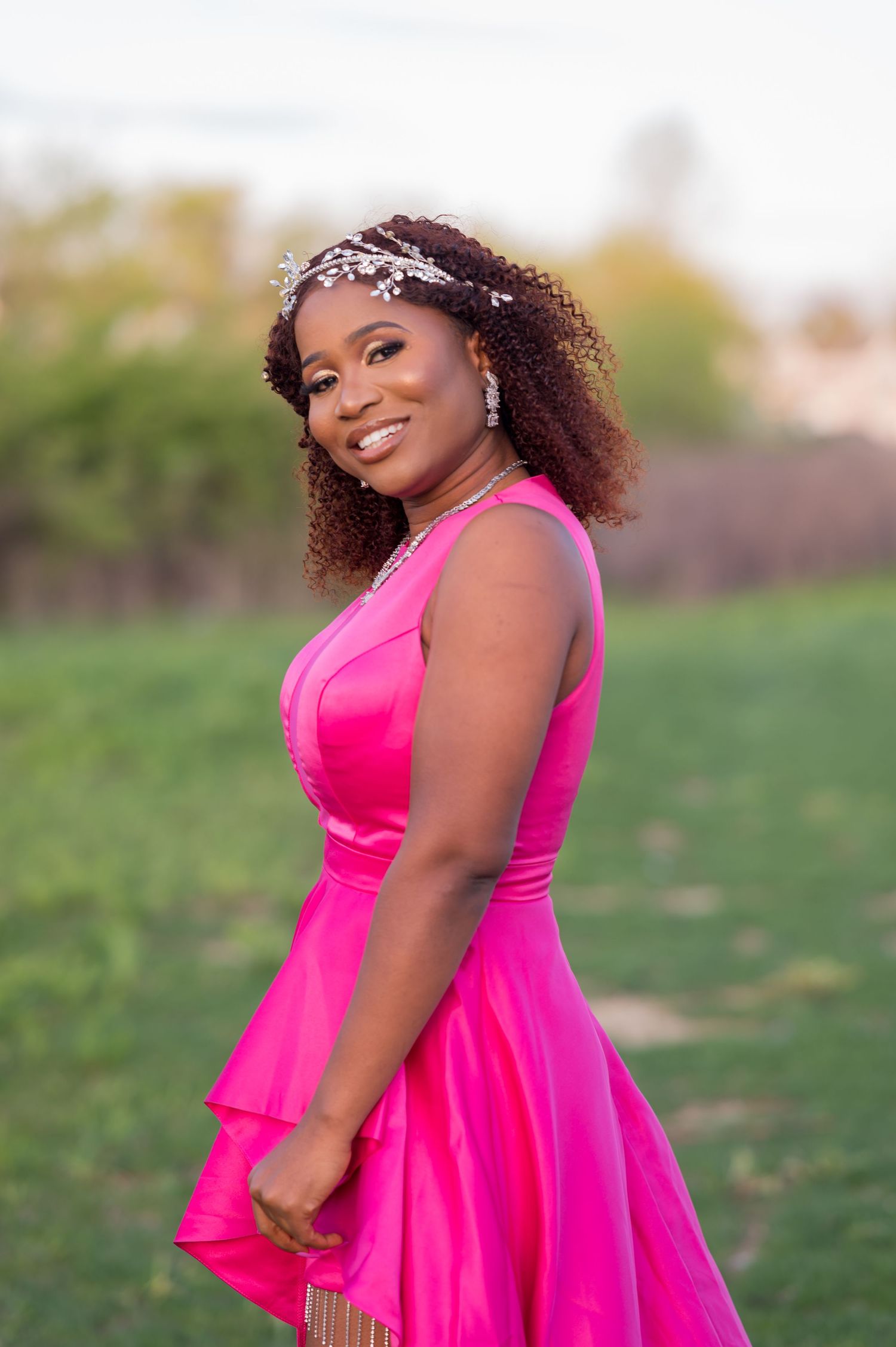 A person in a bright pink dress with curly hair and a sparkly headpiece smiles outdoors.