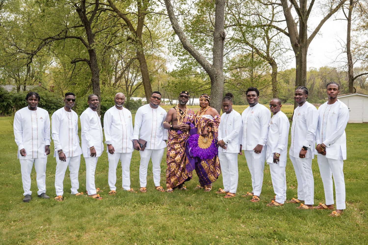 Wedding group photo with white-suited groomsmen and purple-dressed attendants in a park setting.