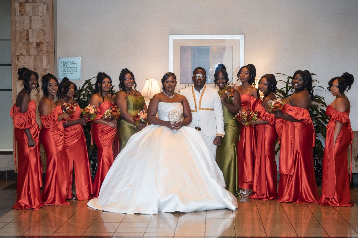 Wedding party members in coral red dresses and olive green suits surround the bride in white.