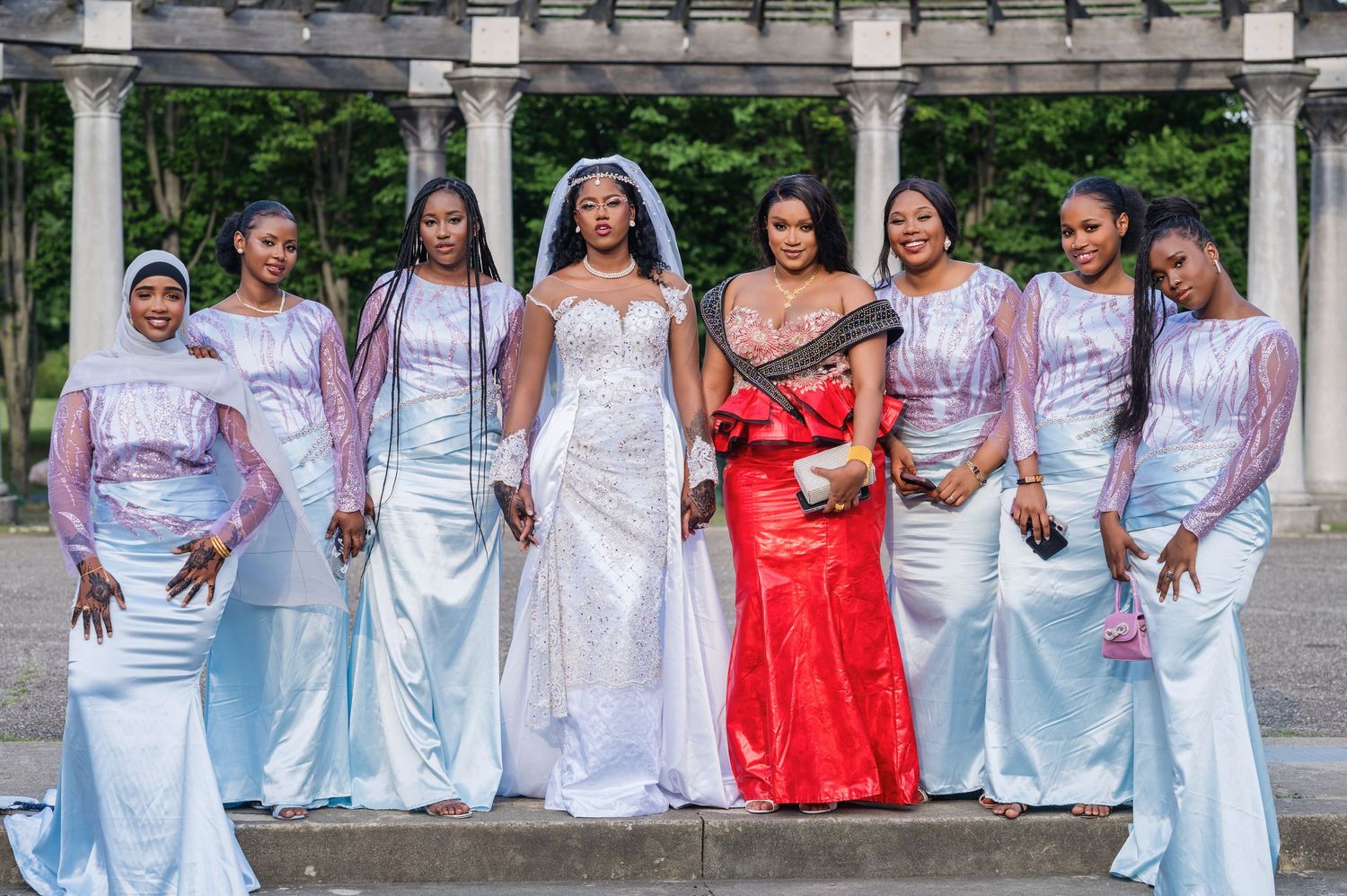 A bridal party in pastel blue dresses and one in red pose together under a white pergola in a garden setting.