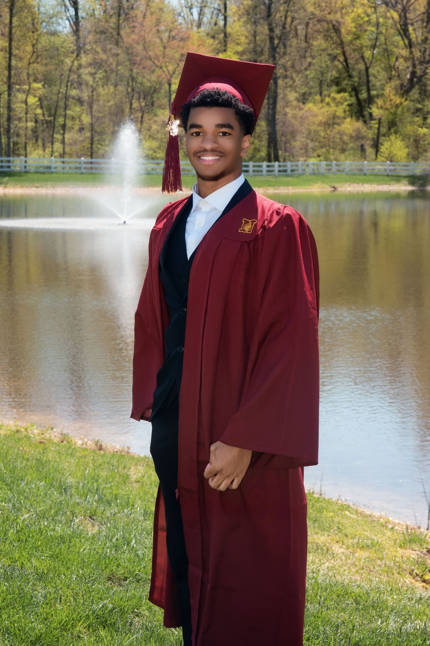 A graduate in a burgundy cap and gown stands smiling on grass near a serene pond with a fountain and autumn trees.