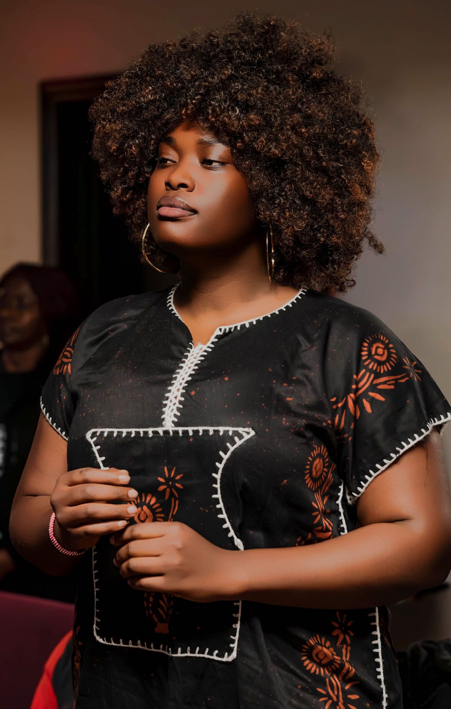A portrait featuring natural curly hair wearing a black embroidered dress with floral details and hoop earrings.