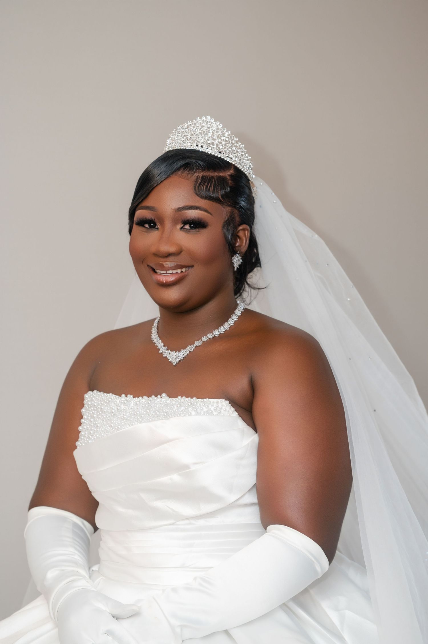 A stunning bridal portrait featuring a white strapless wedding gown, crystal tiara, veil, and elegant pearl necklace.