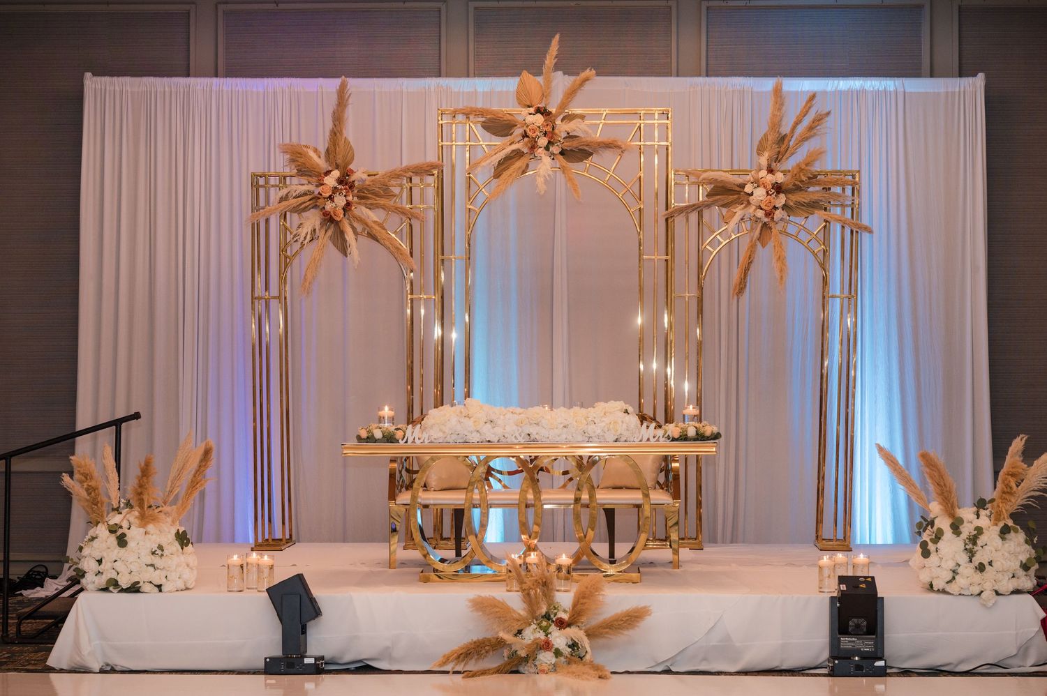 Elegant wedding reception stage with white drapes, golden starburst decorations, and soft blue uplighting.