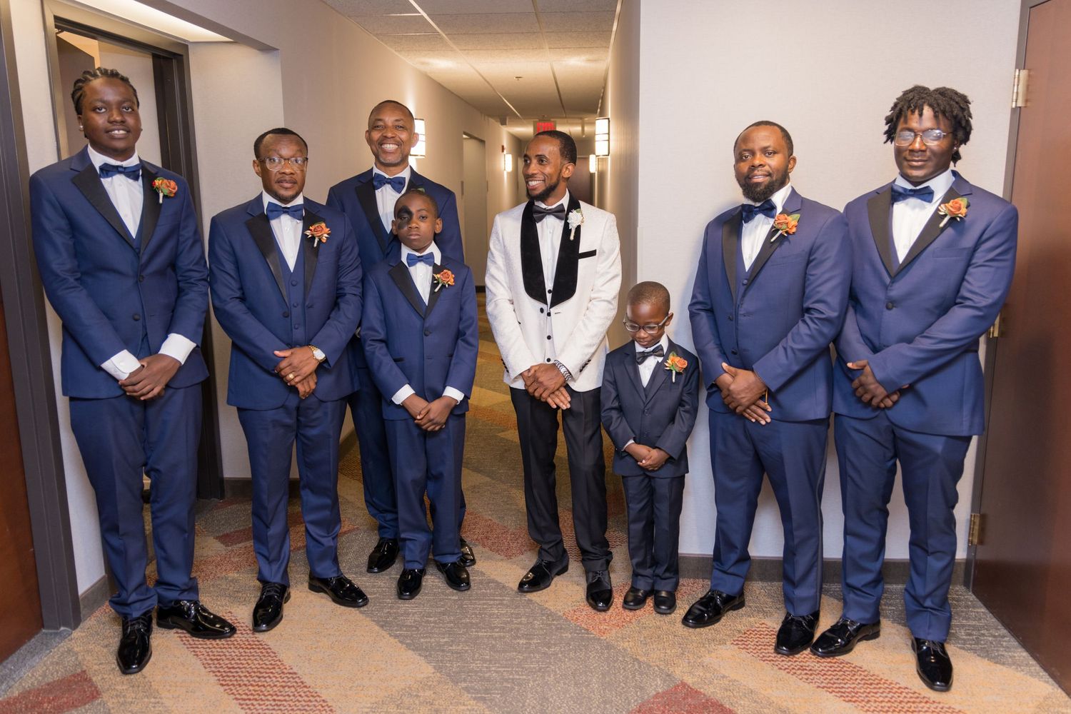 Group of men and boys in matching navy suits and bow ties stand together in a hallway.