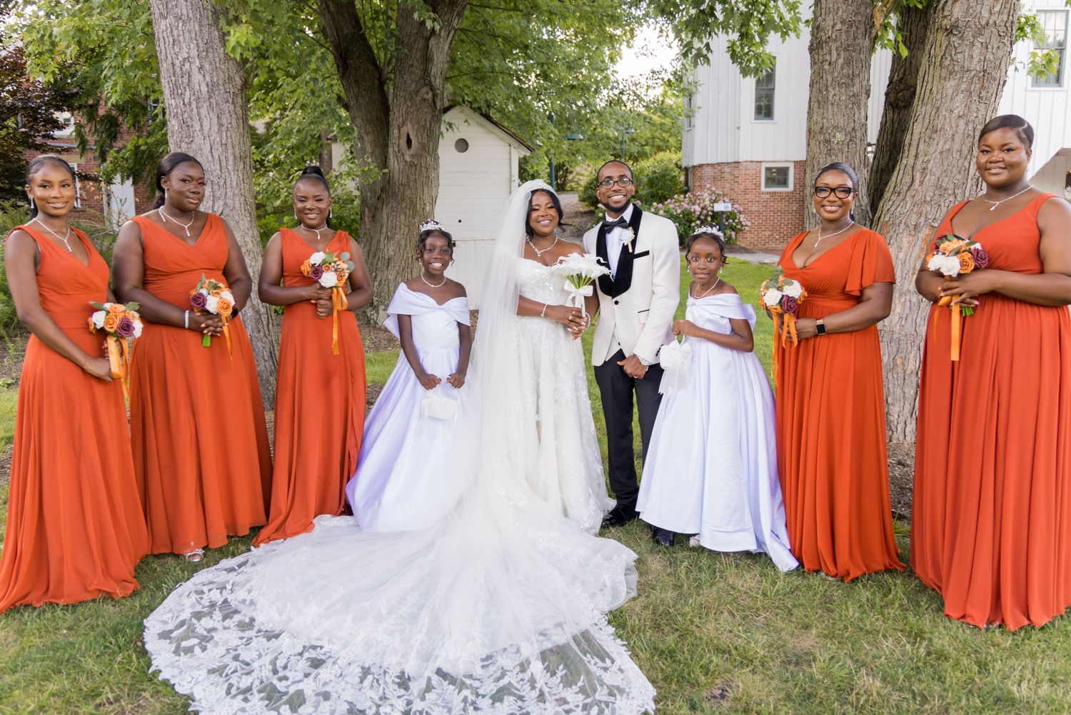 Bridal party with orange bridesmaid dresses and white flower girls pose outdoors with couple.