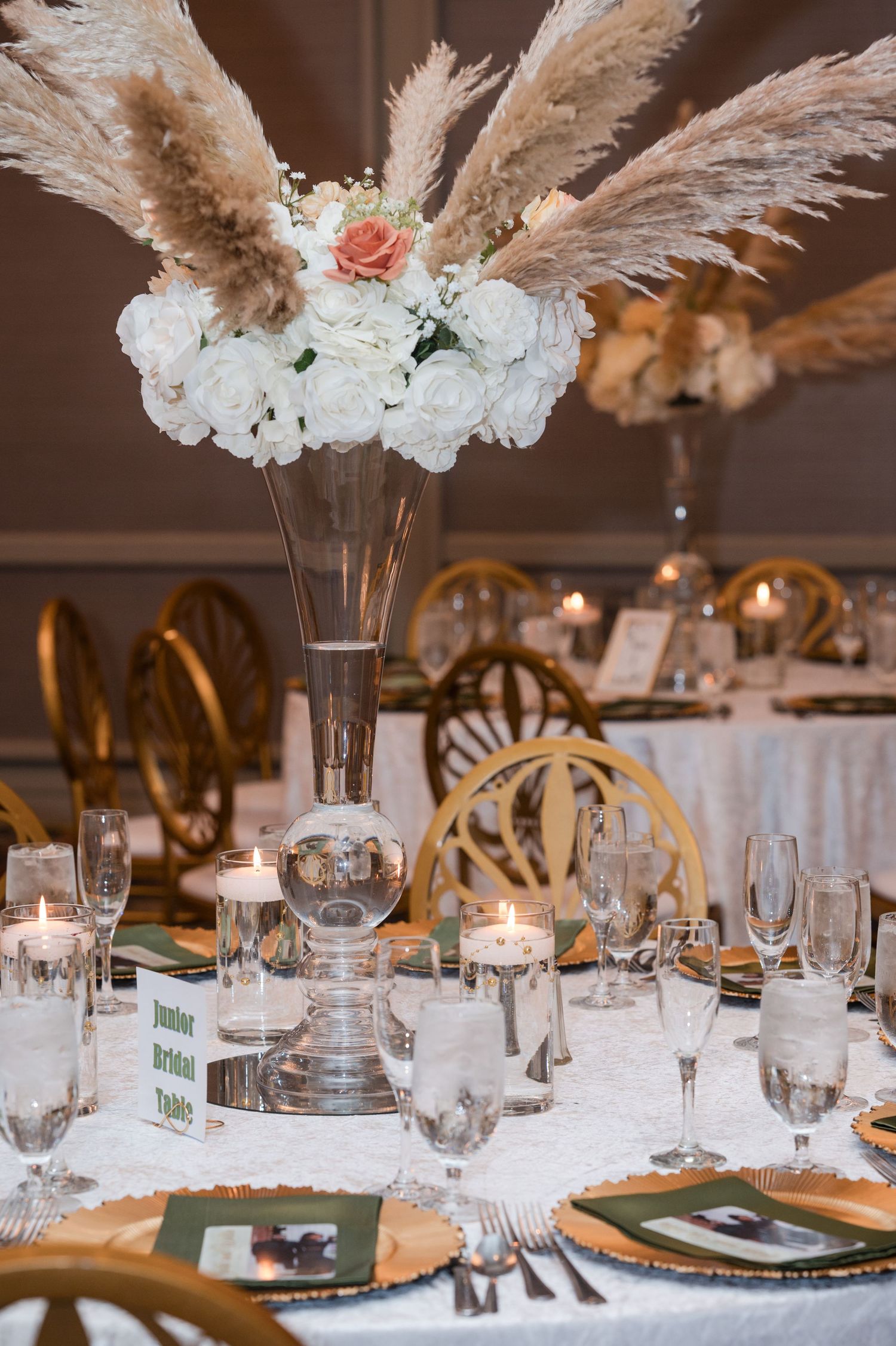 Elegant wedding table setting with white floral centerpiece, pampas grass, gold chargers and glassware for formal reception.