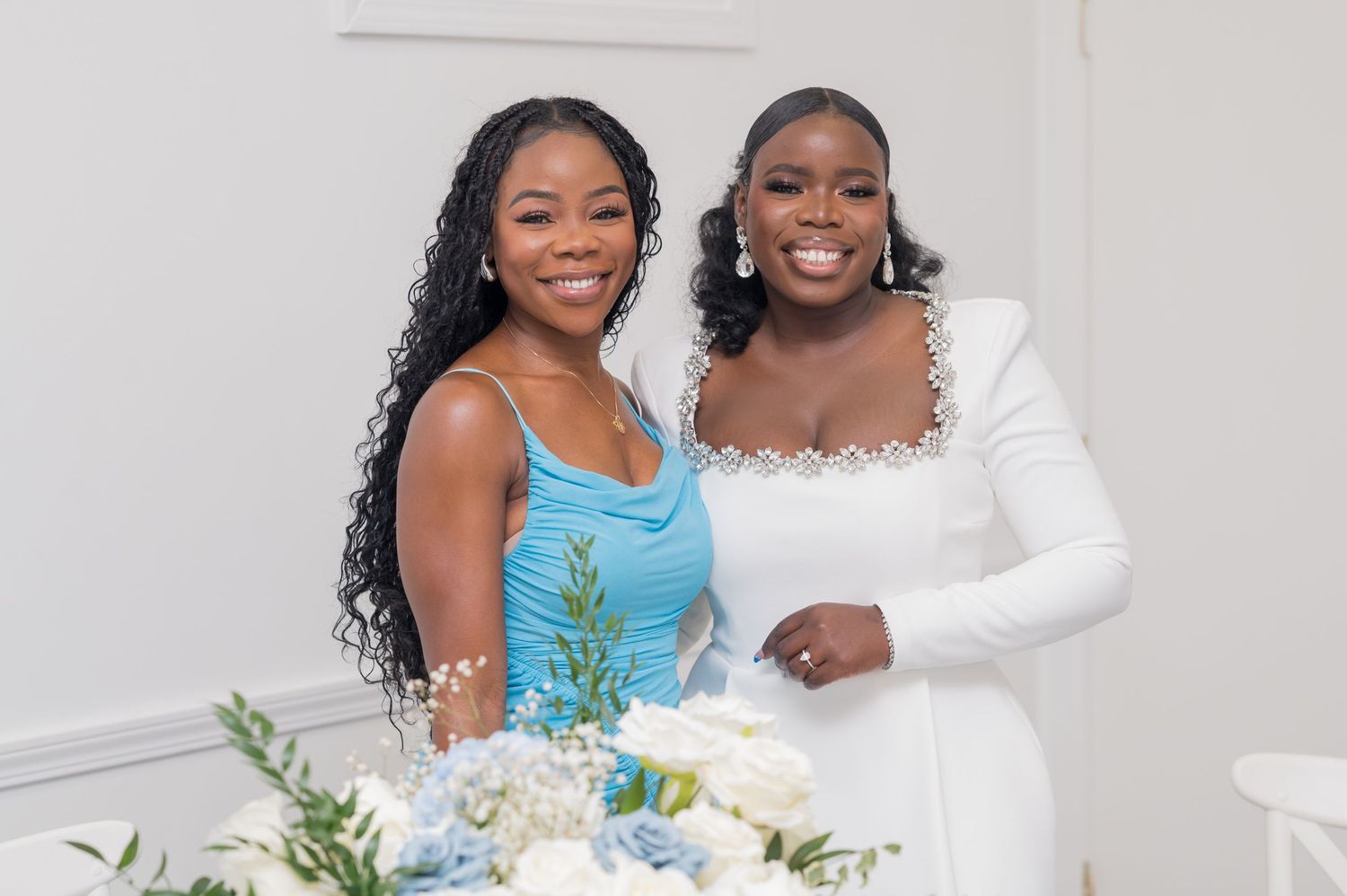 Two women in elegant dresses pose together at a formal event with white and blue floral decorations.