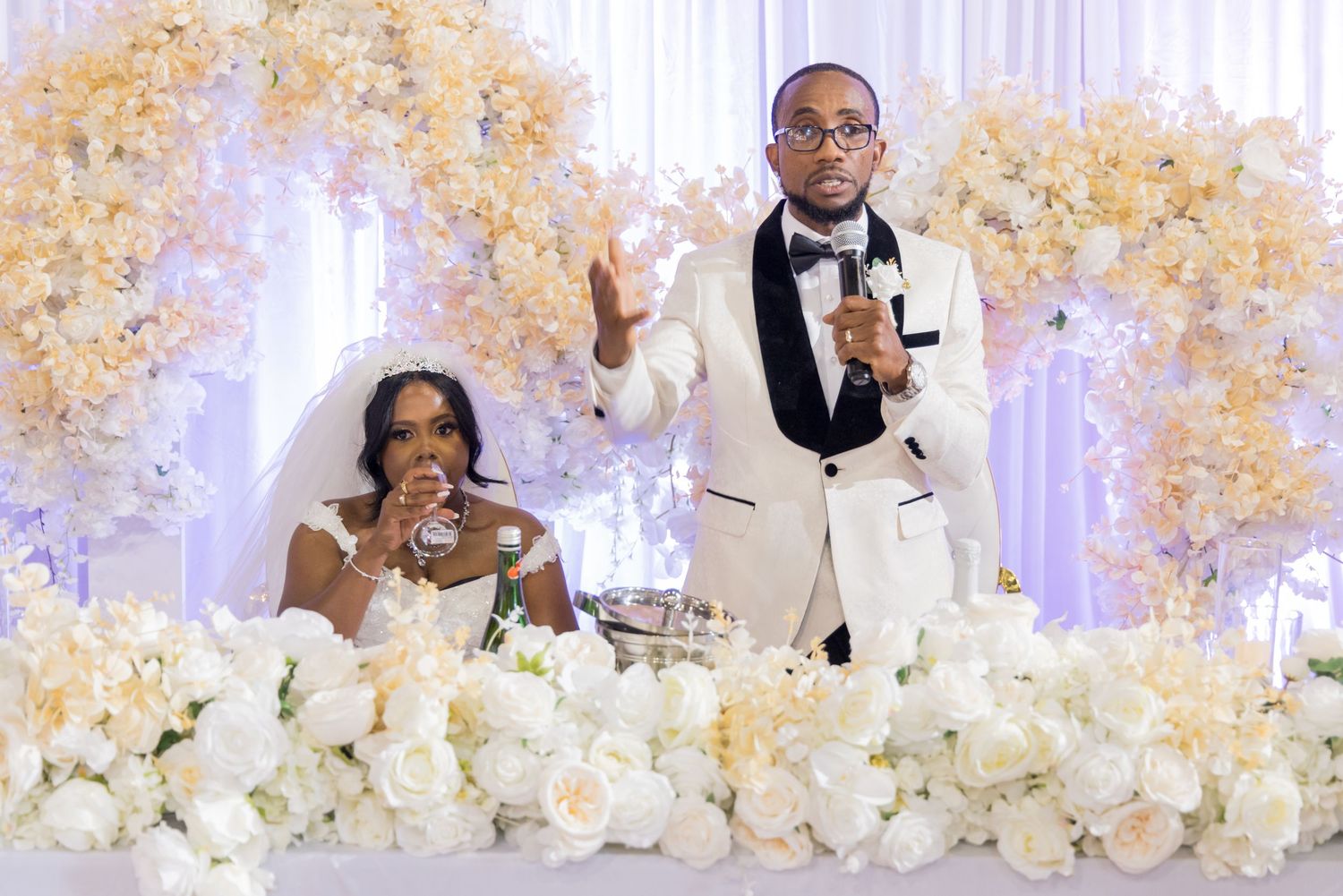 Newlyweds make a toast at their elegant wedding reception surrounded by white and cream floral arrangements and decor.