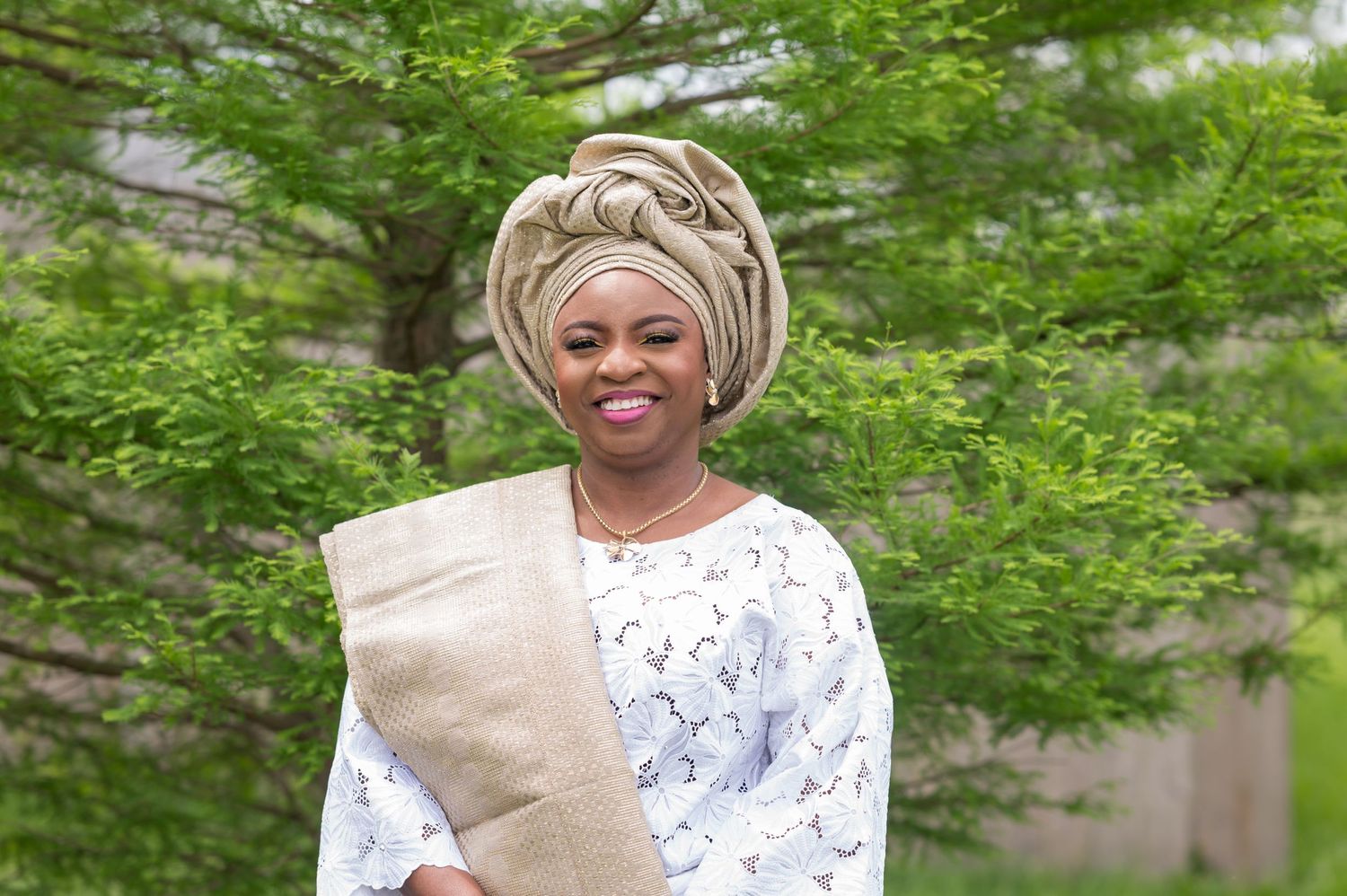 Woman in white lace outfit and elaborate gele headwrap poses outdoors with beige accessory.