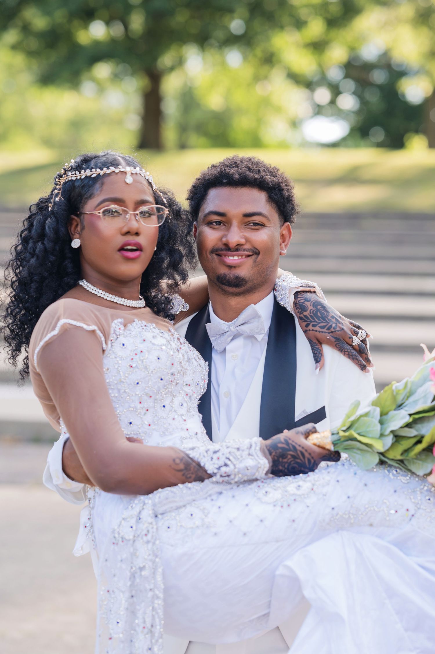 A happy couple poses on their wedding day in warm sunlight, with the bride wearing a sparkly white dress and lace sleeves.