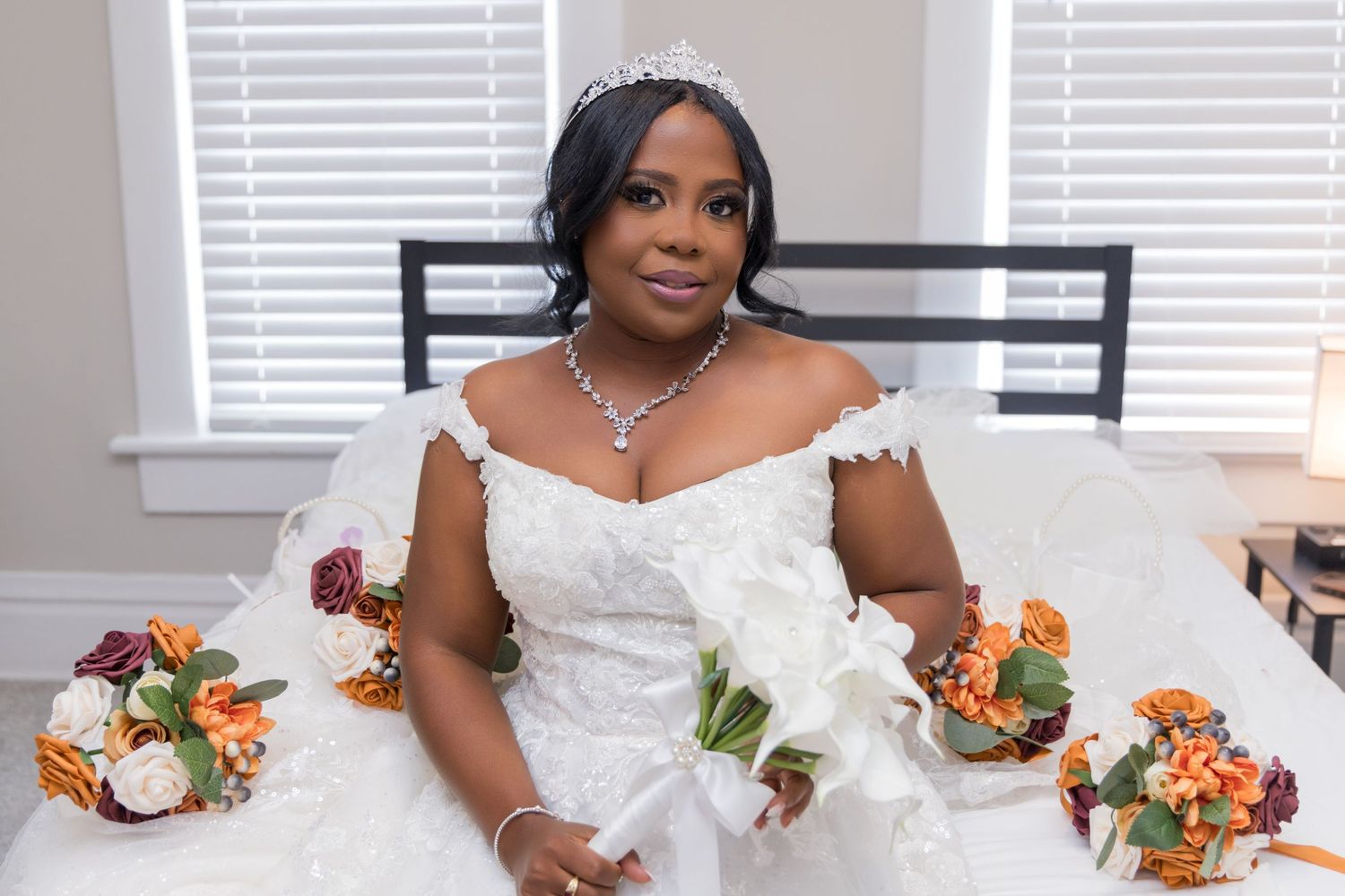 A bride in white lace dress poses with orange and white floral bouquets against a light background.