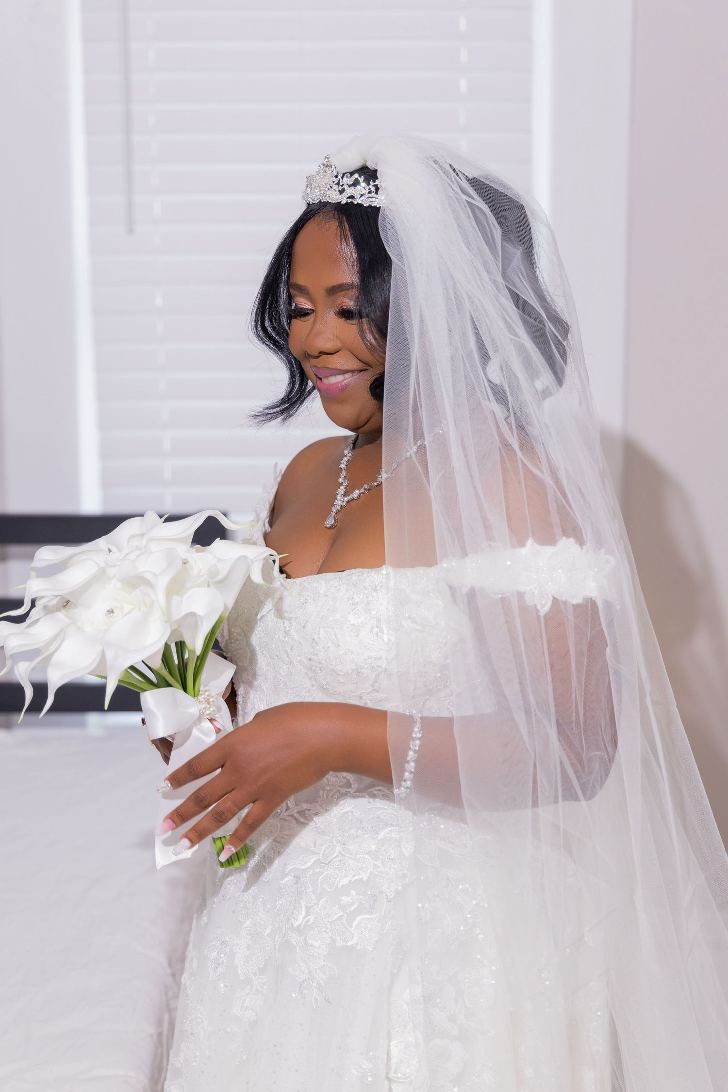 A stunning bride in a traditional white wedding dress and veil smiles while holding a white calla lily bouquet.