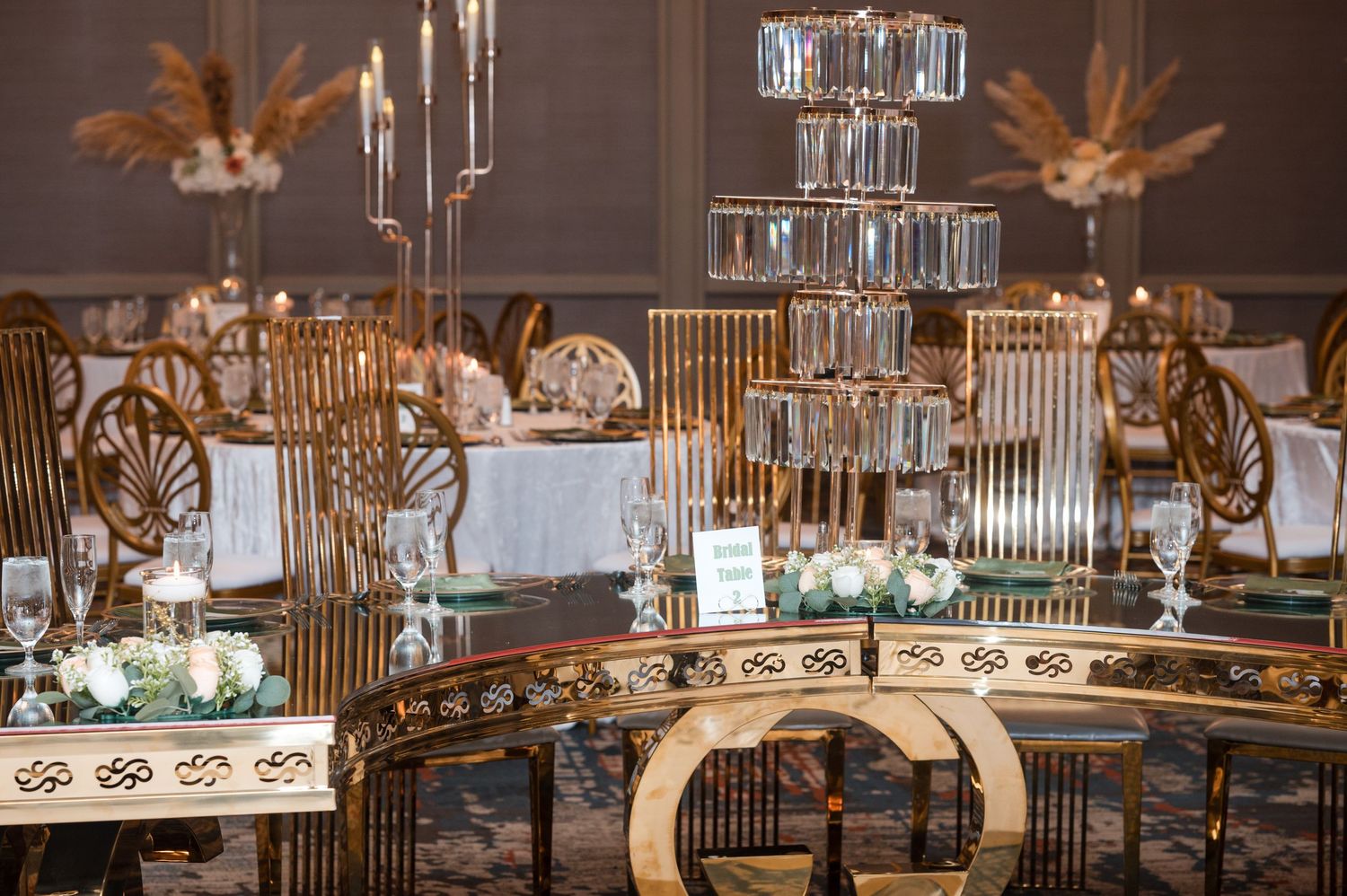 Elegant wedding reception table with gold chairs and crystal candlesticks surrounding a tall tiered glass centerpiece.