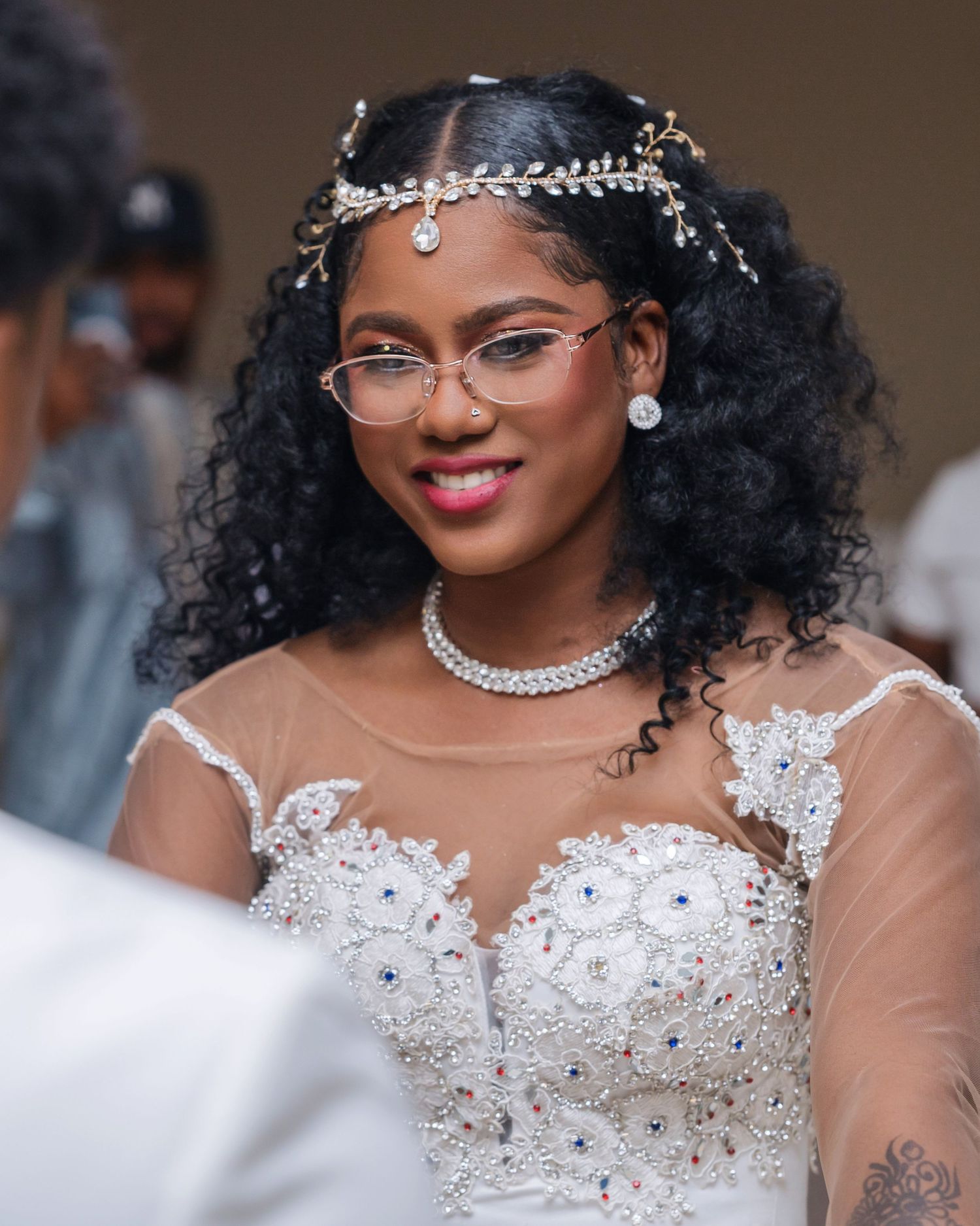 A bride wearing an elegant sheer lace wedding dress with crystal embellishments, glasses and a delicate headpiece.