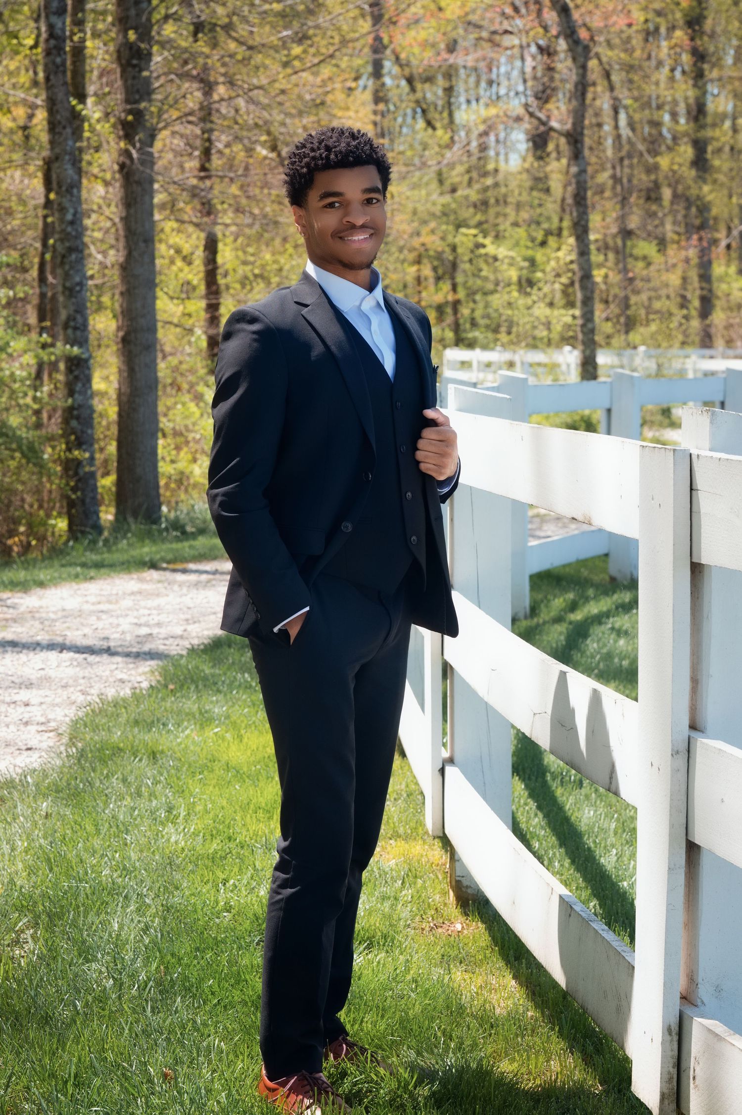 A person in a formal black suit poses against a white fence on a sunny spring day with autumn trees in the background.