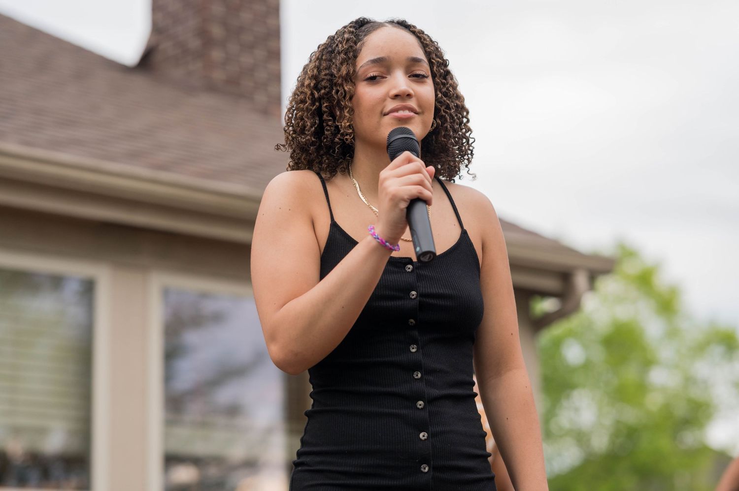 A performer in a black dress sings into a microphone at an outdoor event.
