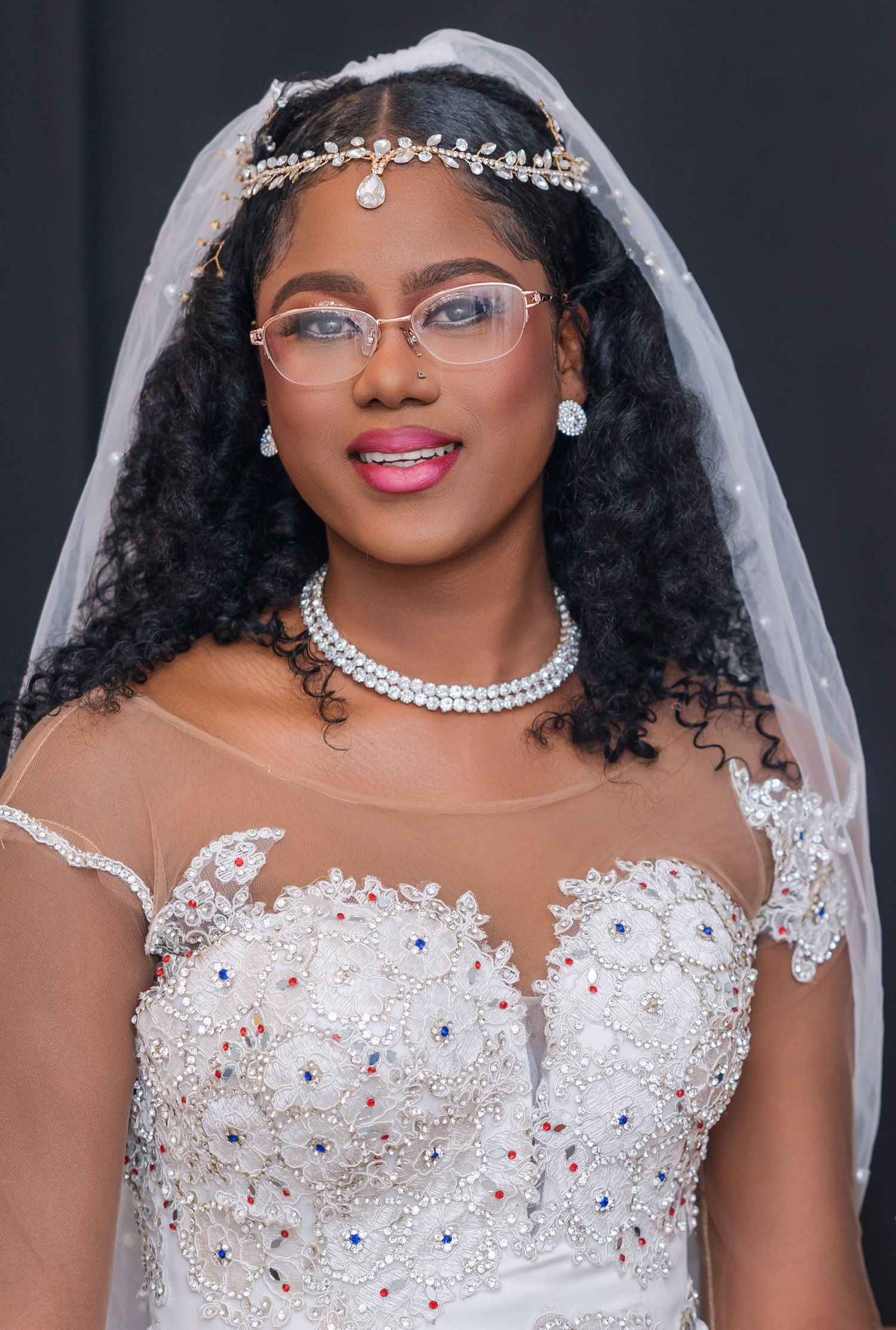 A bride wearing an embellished white lace wedding gown with glasses, crystal headpiece and veil smiles at the camera.
