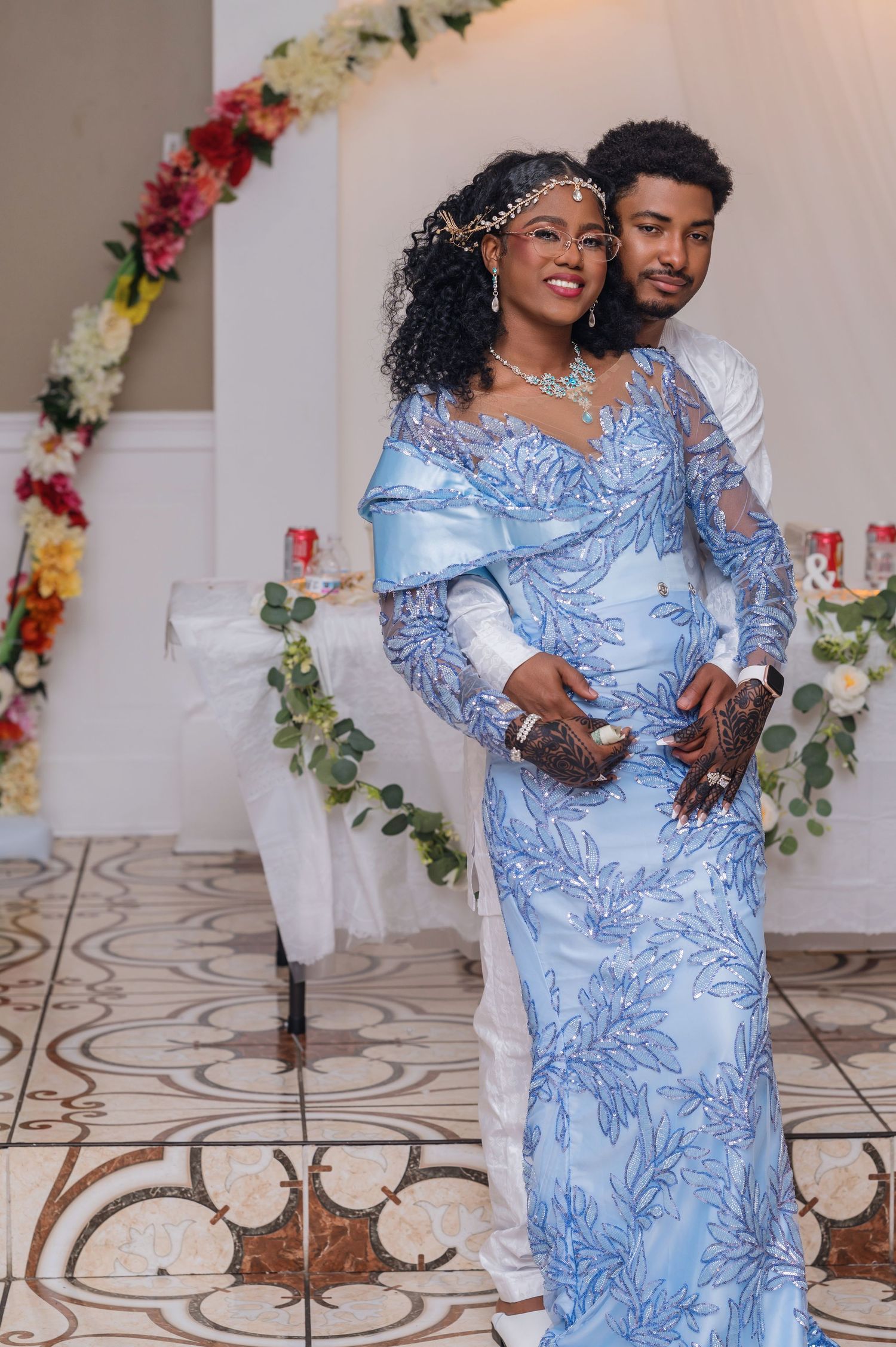 A couple poses together in front of a floral arch with the woman wearing a flowing light blue long-sleeved gown.