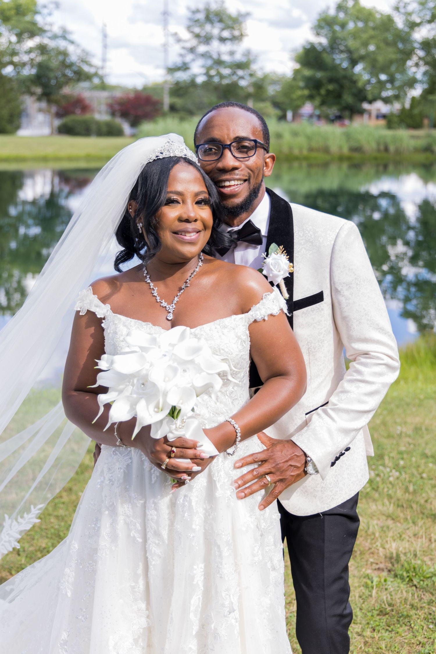 A wedding couple poses by a lake with the bride in a white lace gown and the groom in an ivory tuxedo jacket.