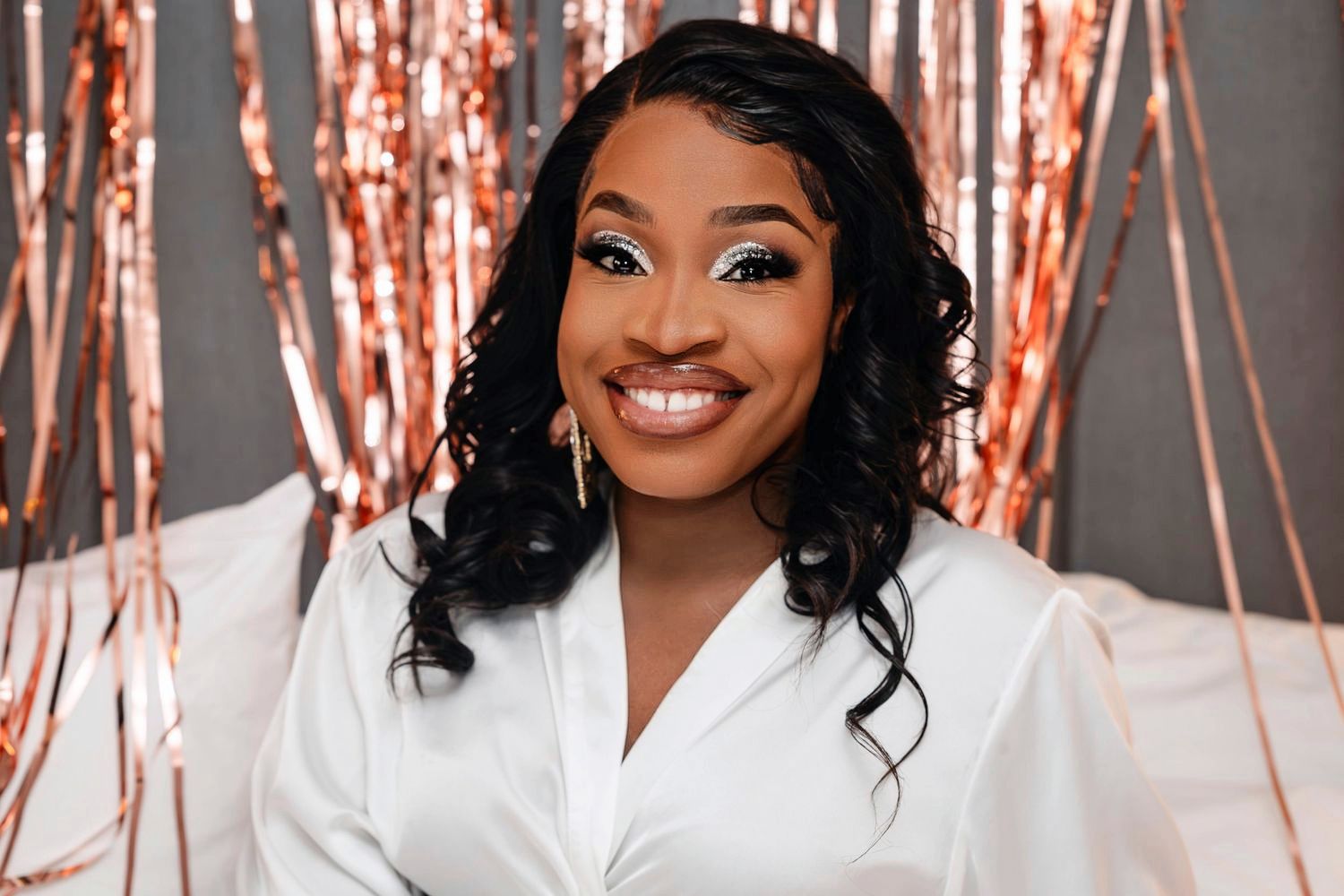 A woman wearing a white blazer smiles warmly against a backdrop with rose gold decorative elements.