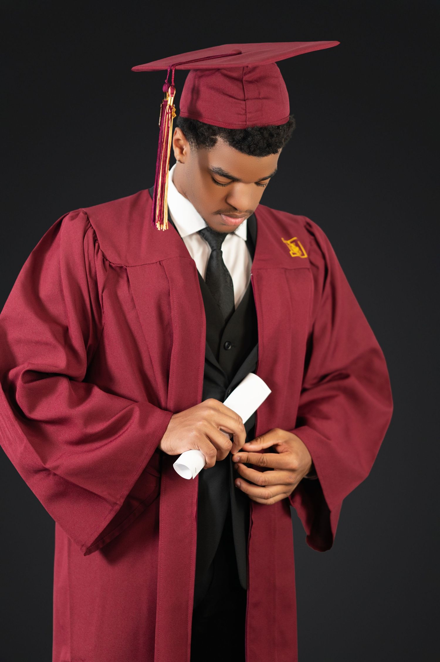 Graduate in maroon cap and gown holding diploma while looking down against dark background.