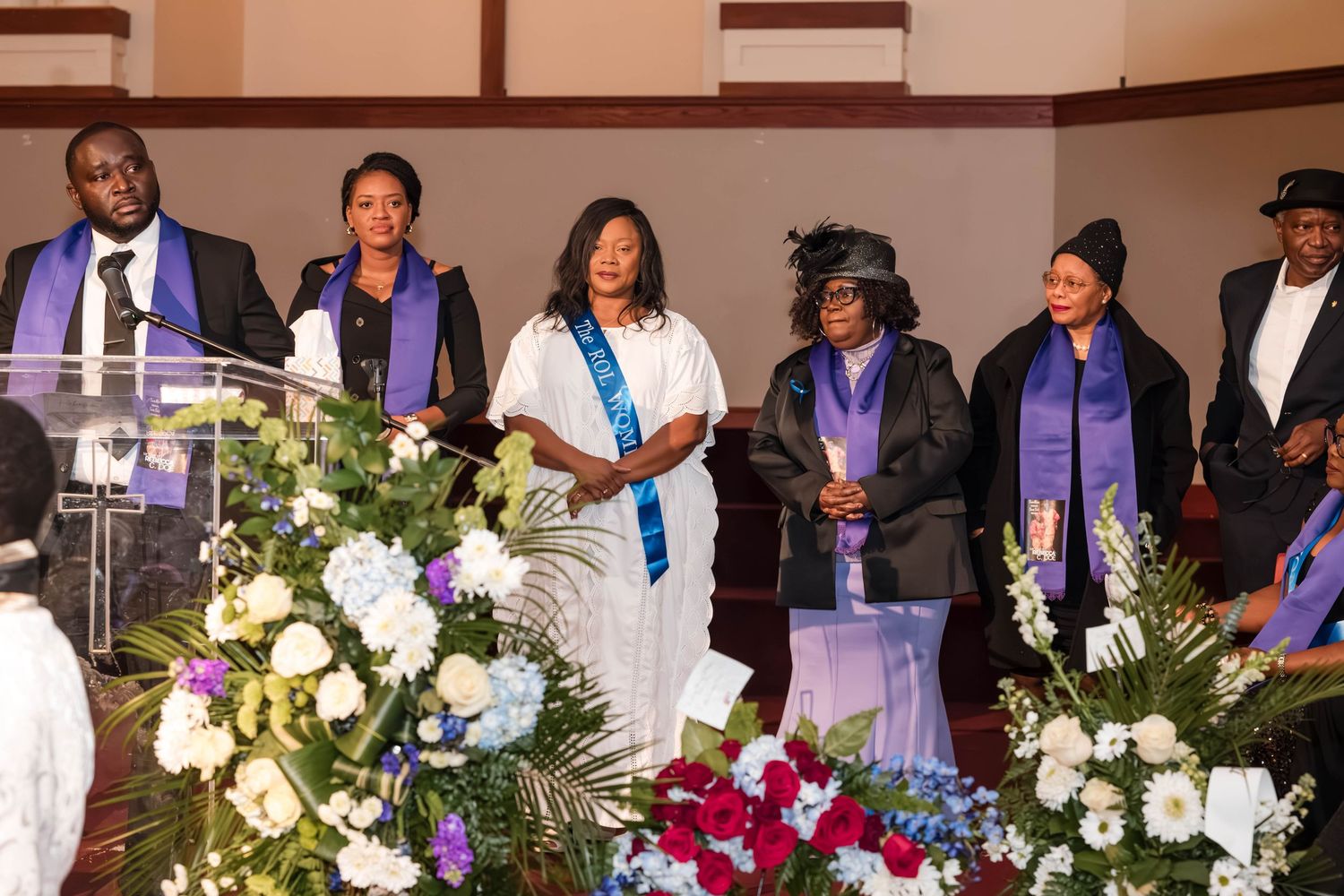 Group standing together at ceremonial event with purple robes and white floral arrangements on display.
