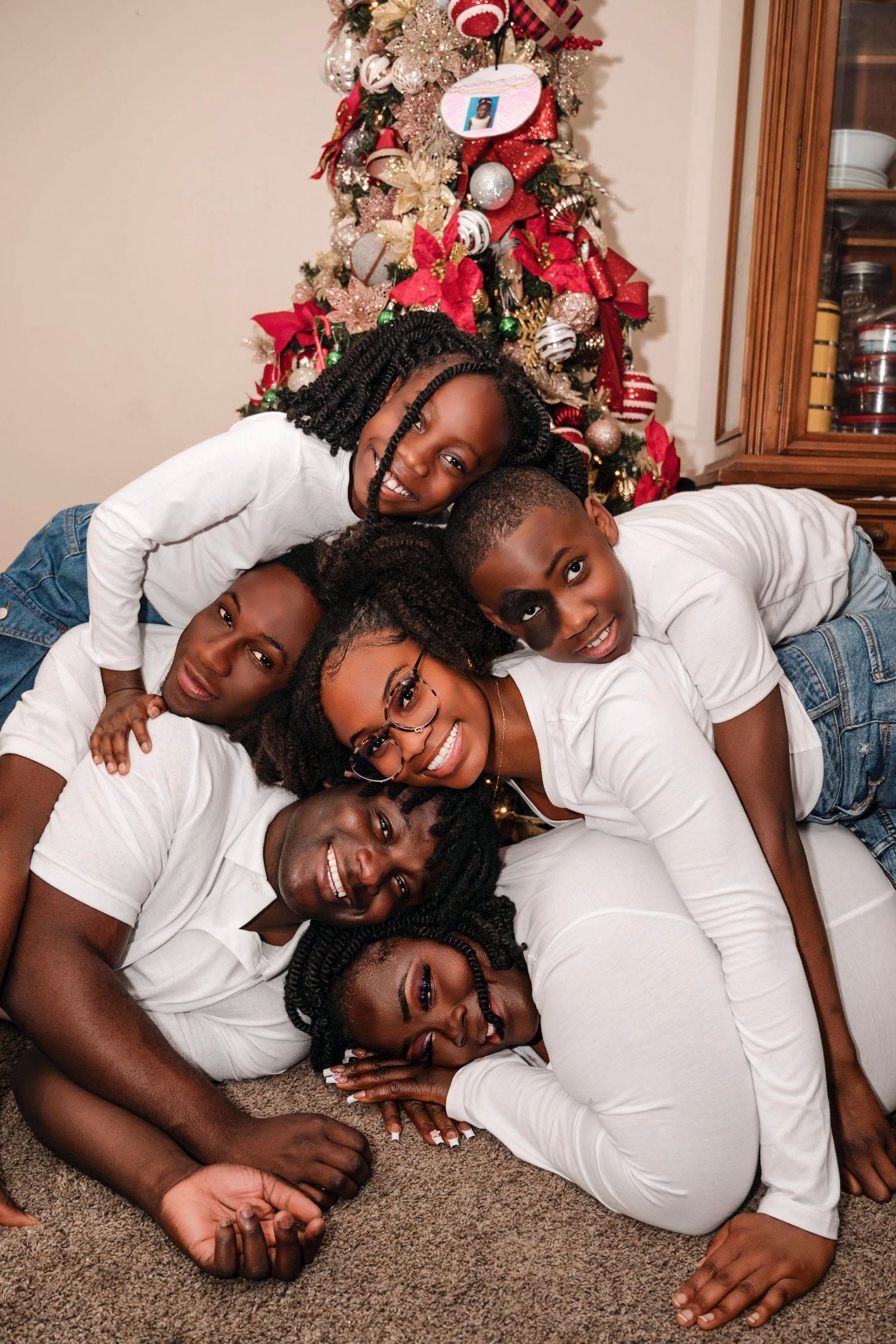 A group in matching white shirts poses together on the floor in front of a decorated Christmas tree with red ornaments.