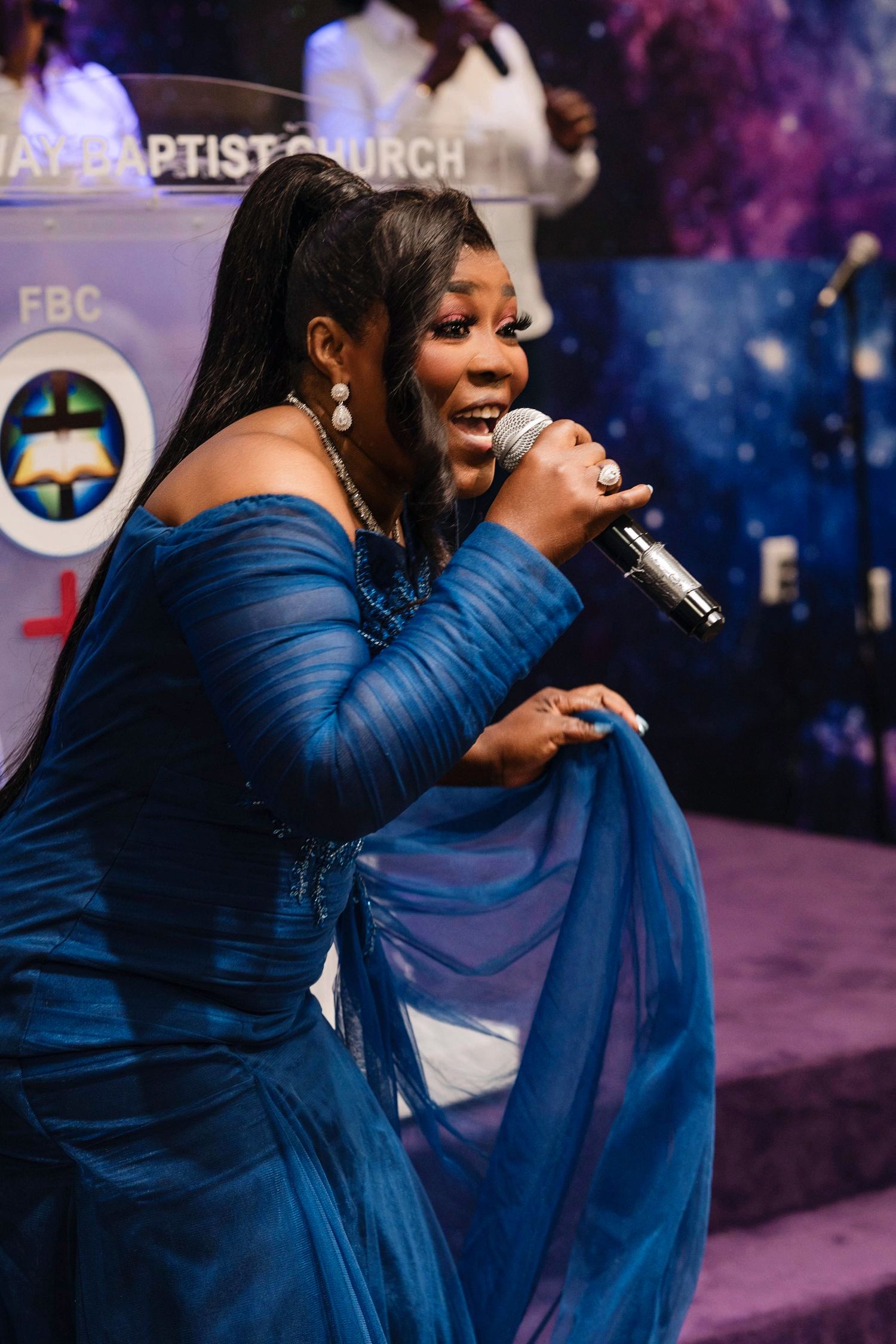 A singer performs passionately in a royal blue gown during a church service with colorful stage lighting and a purple backdrop.