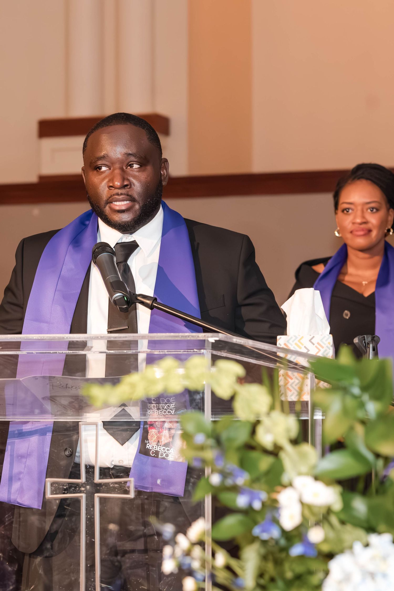 A speaker wearing a purple stole addresses an audience from behind a clear podium with flowers.
