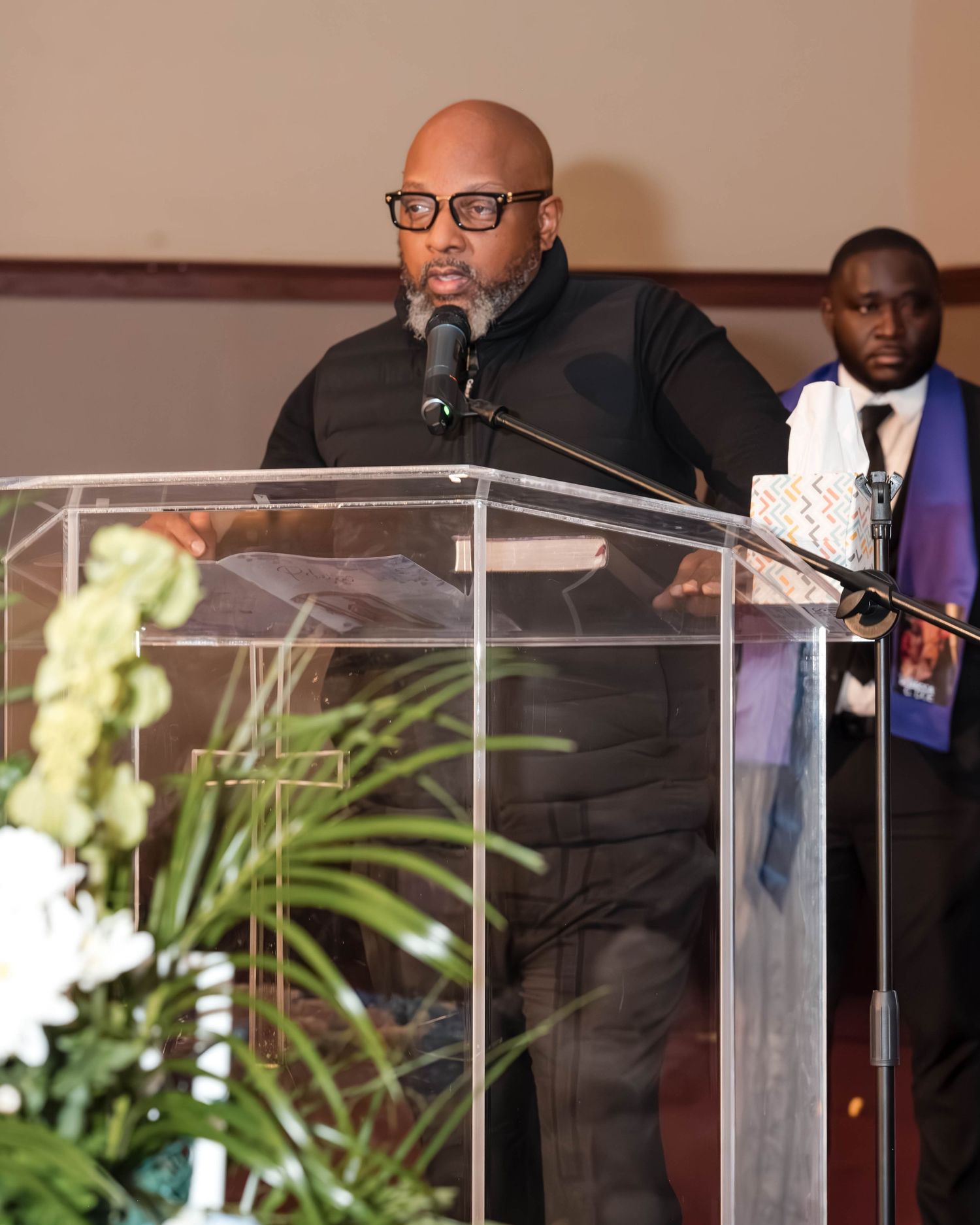 Speaker at podium wearing glasses and black jacket delivers remarks from clear acrylic lectern with floral arrangements.