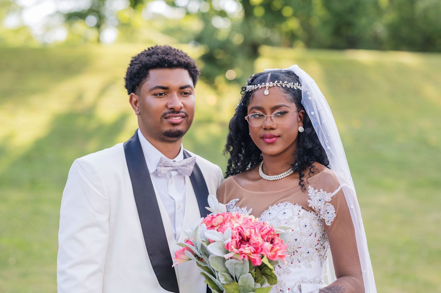 Happy newlyweds smile in white formal wedding attire while holding pink bouquet in sunny outdoor garden setting.