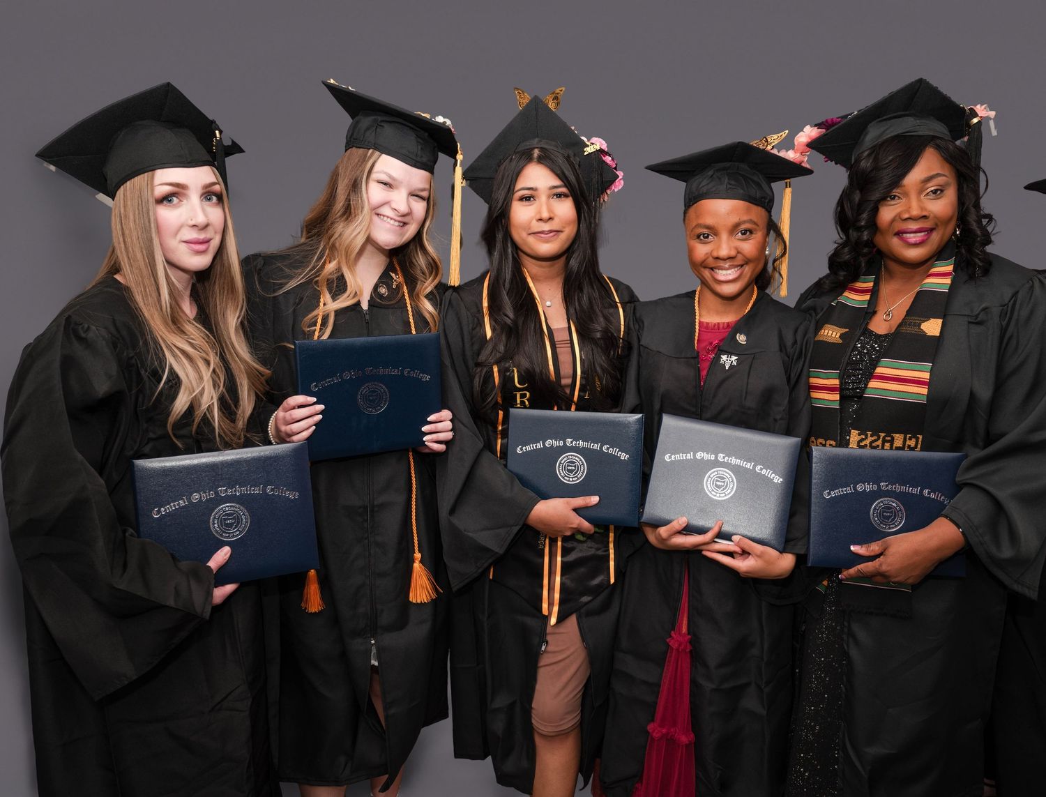 Five graduates in black caps and gowns proudly display their diplomas while posing for a group photo.