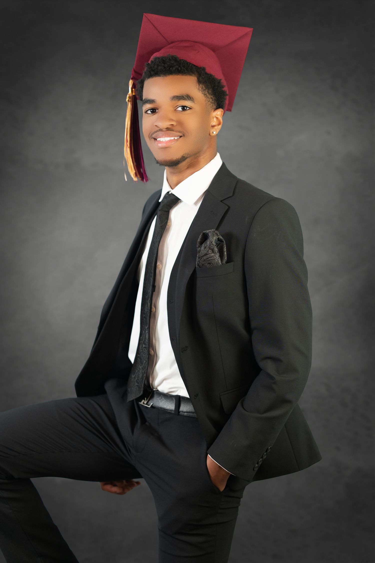 Graduation portrait of a graduate in a burgundy cap and black suit standing against a dark gray backdrop.