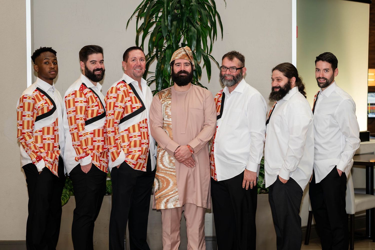 A lineup of groomsmen wearing white suits and orange patterned jackets stand together at a wedding event.