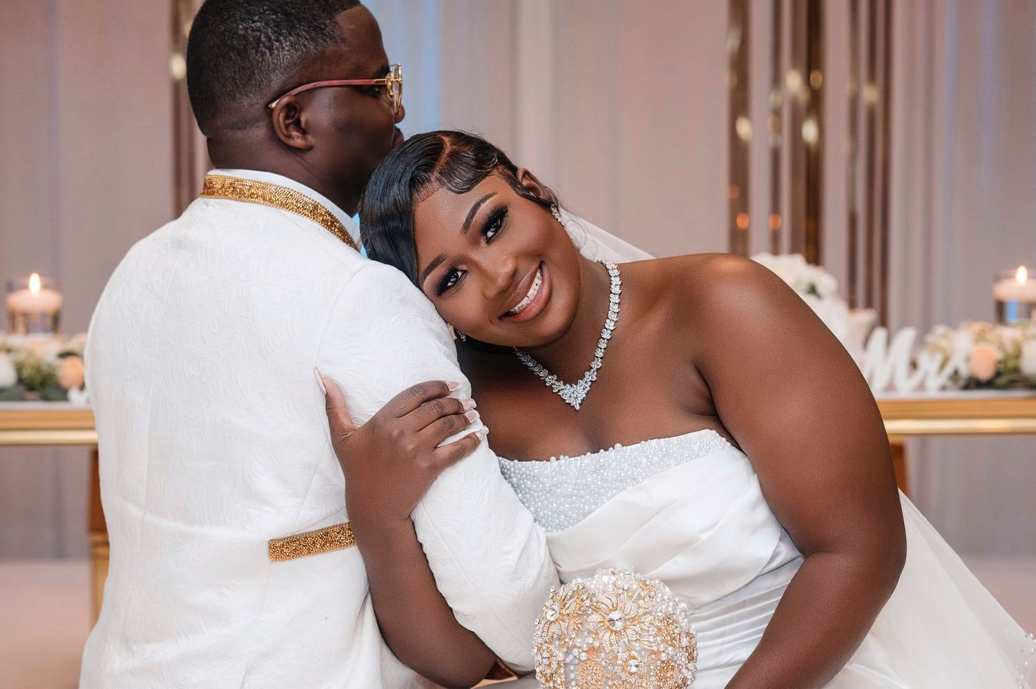 A smiling couple embraces in formal wedding attire against an elegant indoor reception venue backdrop.