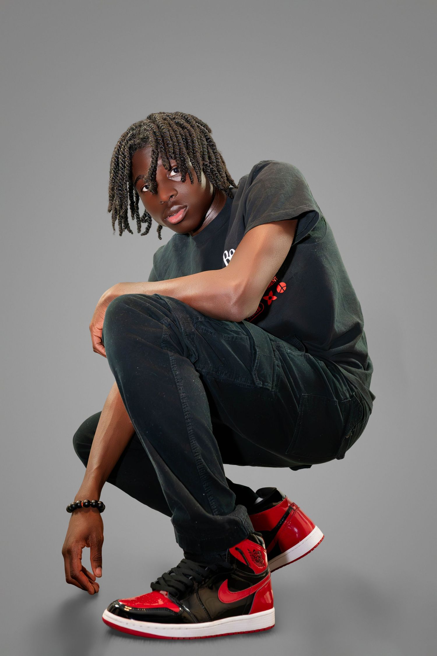 Person in dark clothing crouches wearing red and black Air Jordan sneakers against grey studio background.