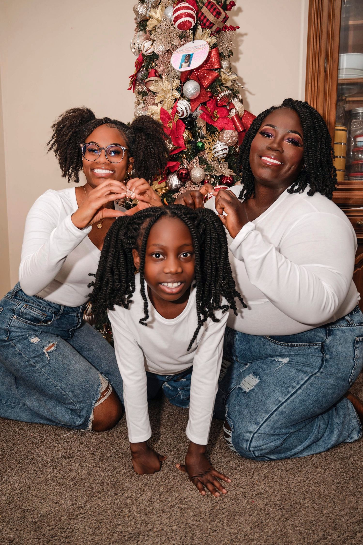 Three people in matching white tops and jeans pose together in front of a decorated Christmas tree at home.