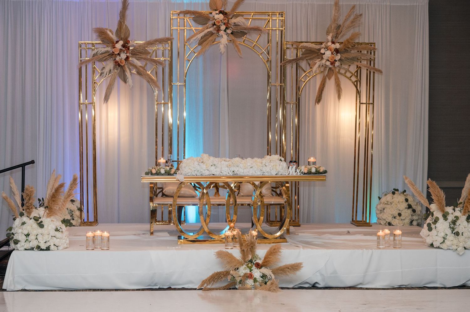 Elegant gold and white wedding reception stage with ornate mirrors, pampas grass decor, and floral arrangements.